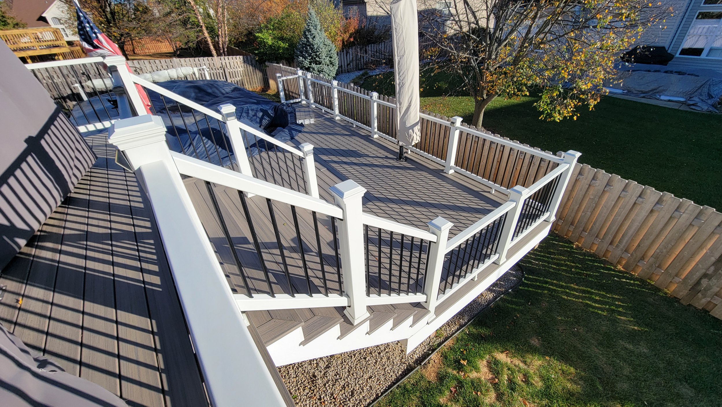 A wooden deck with white and black railing surrounds a backyard. There is a staircase leading down from the deck, an outdoor umbrella, and a blue tarp covering an object. The yard has a tree, grass, and a wooden fence.