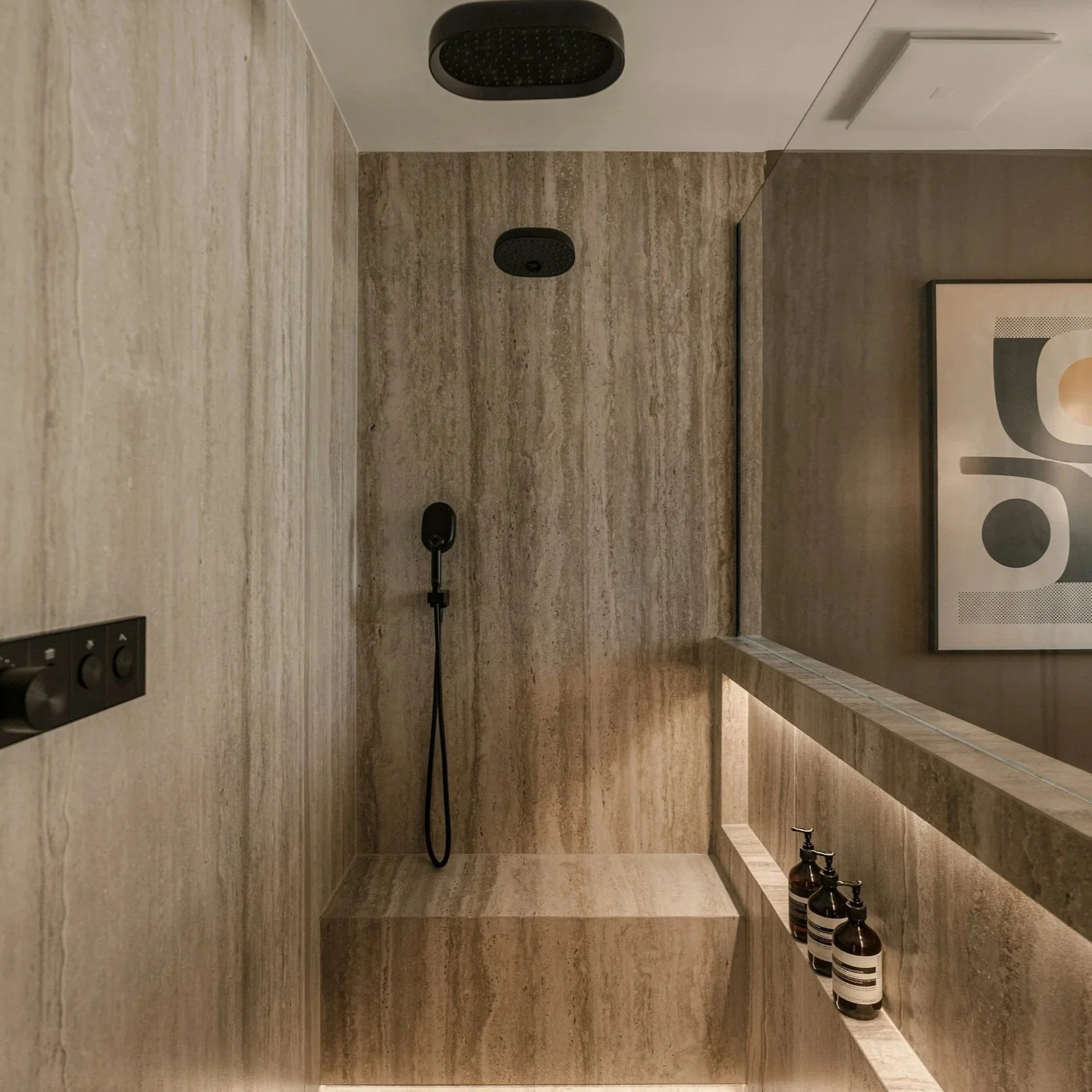 Modern hotel shower with beige marble walls, black shower fixtures, and bottles of shampoo and body wash on a ledge.
