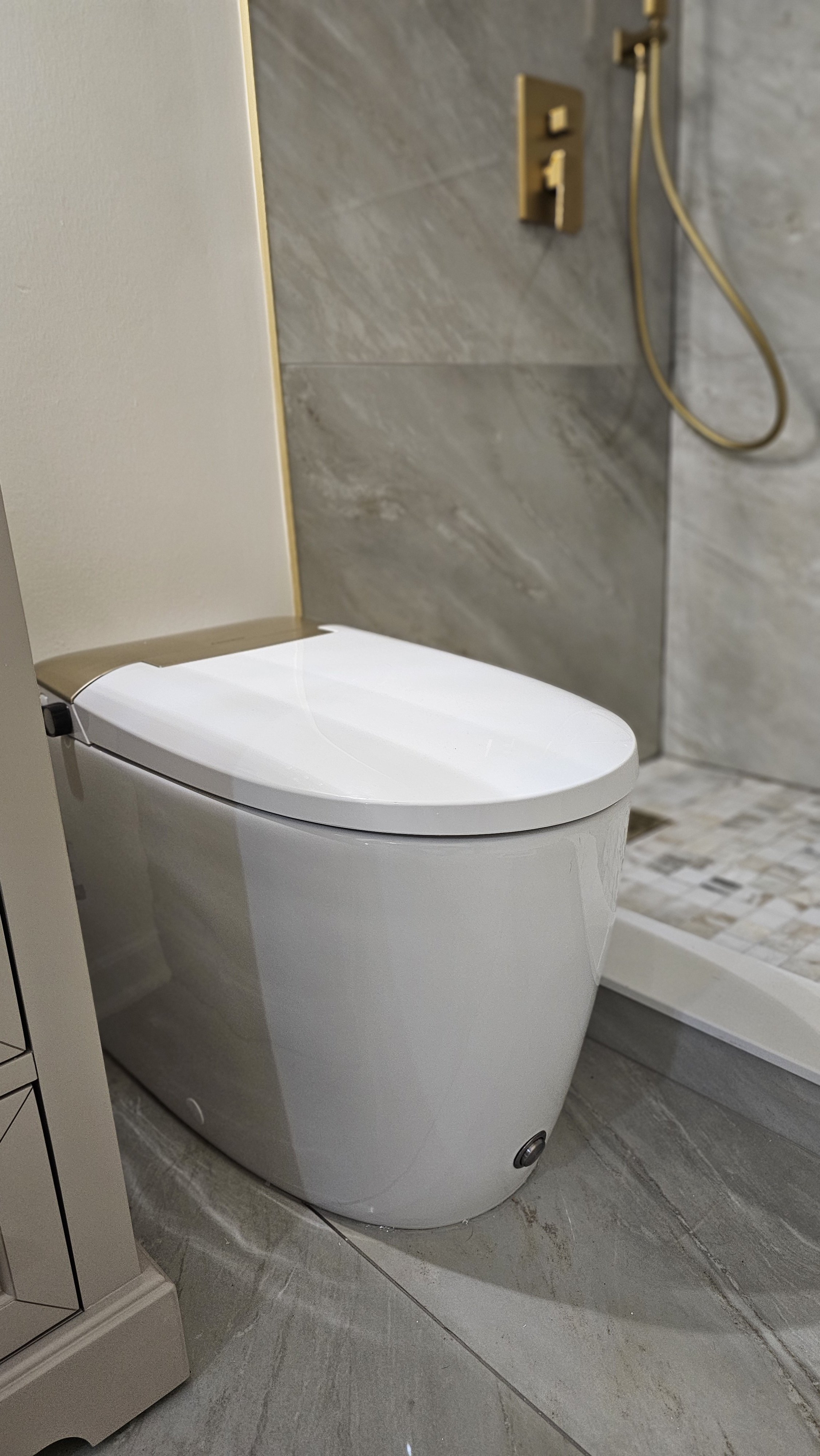 Modern white toilet in a bathroom, with a shower area featuring a gold-colored handheld showerhead and faucet on a gray tiled wall.