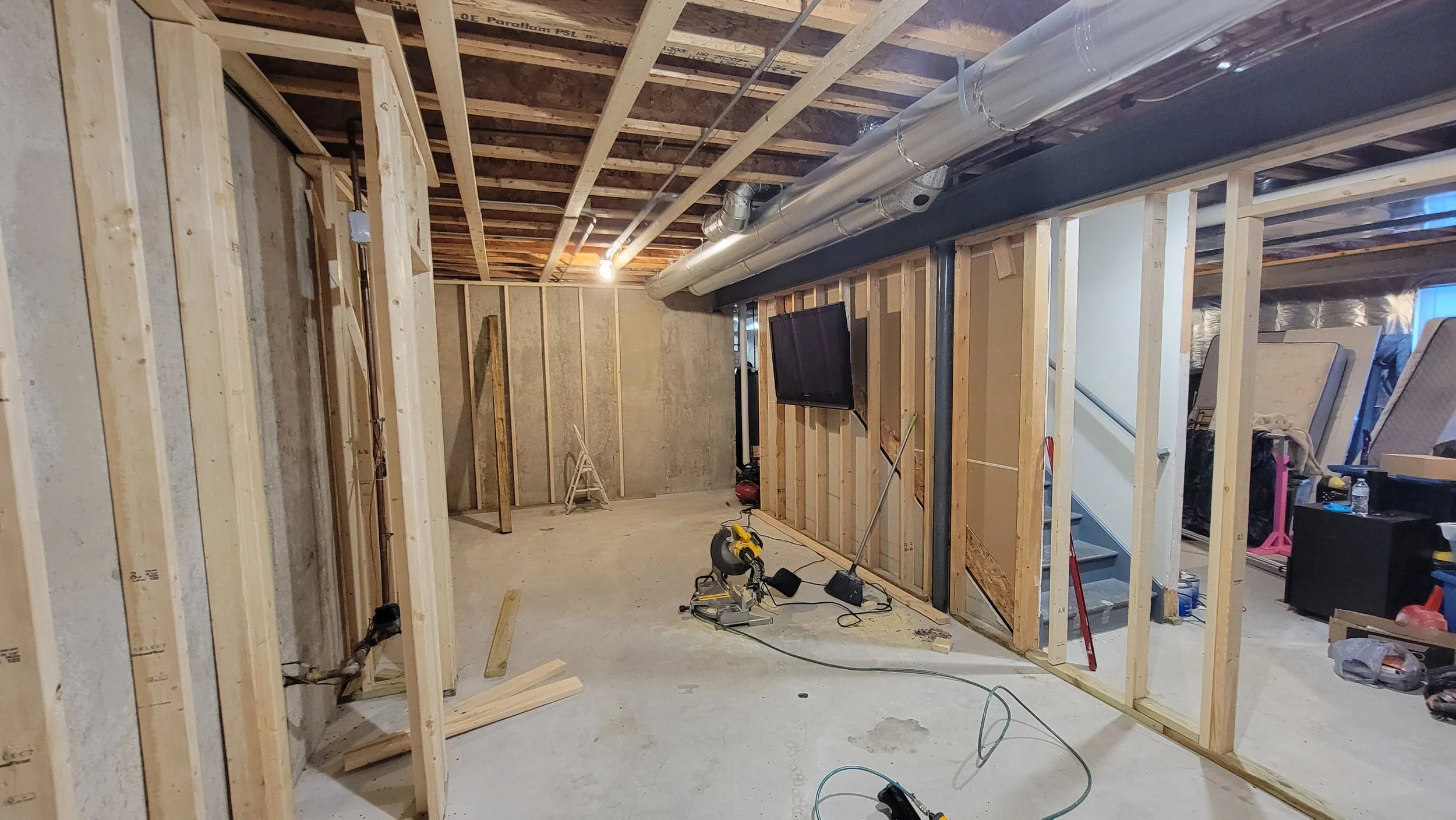 Underground basement construction with exposed wooden framing, drywall, and visible plumbing and ductwork, including a mounted television and construction tools on the floor.