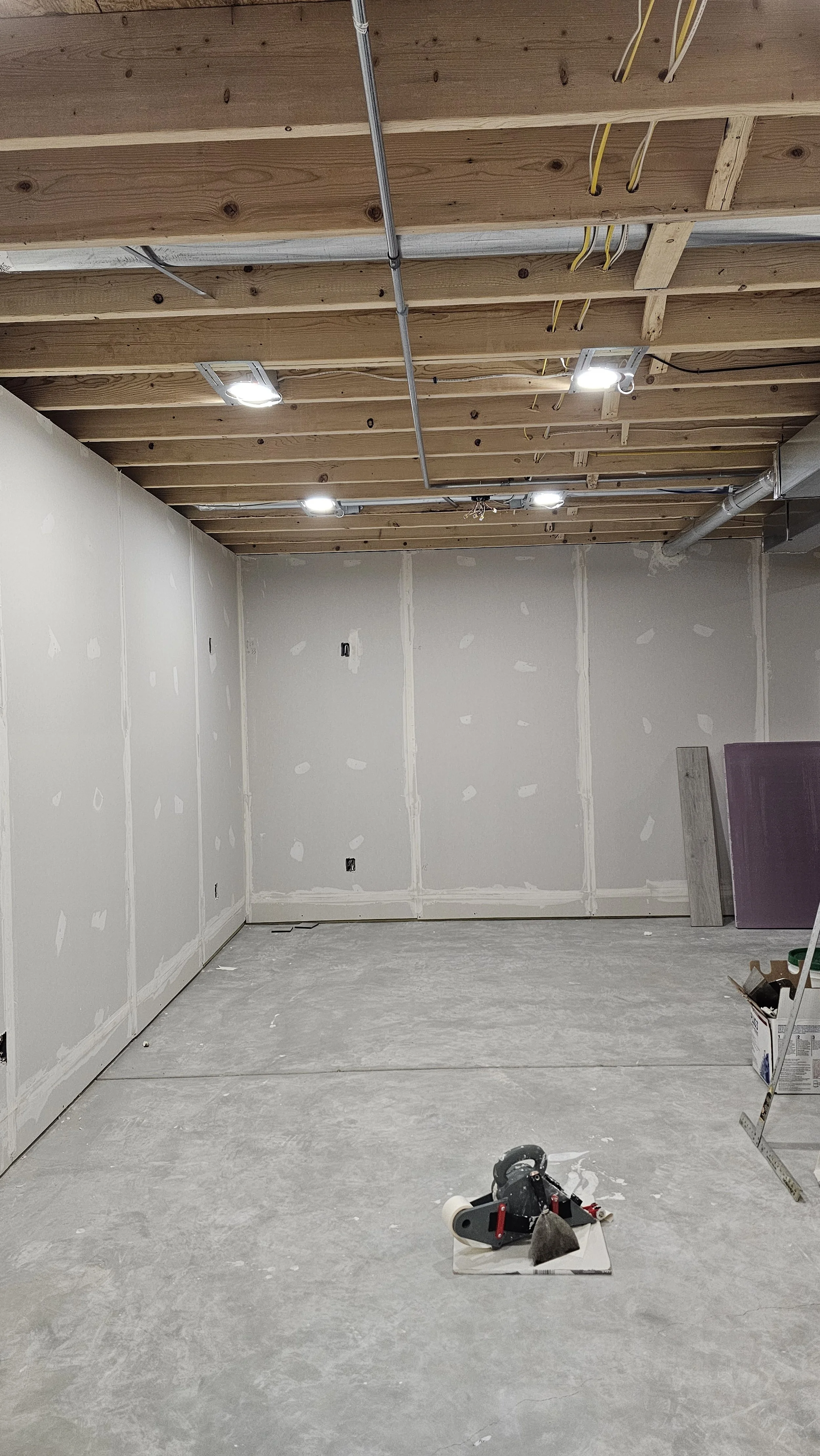 An unfinished room with drywall walls, a wooden ceiling with electrical wiring and lighting, and a concrete floor. Construction tools and materials are present, including a scaffold on the right side.