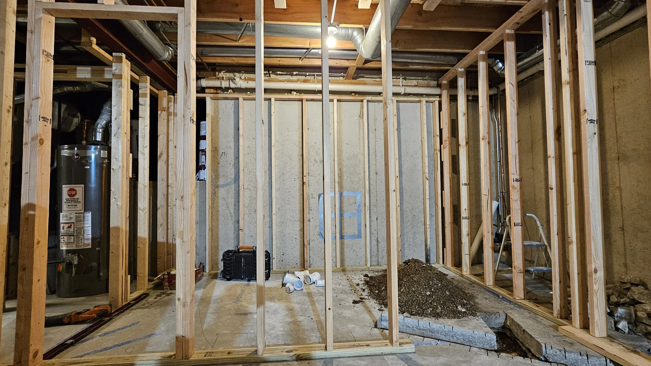 Interior view of a building under construction with wooden framing, exposed pipes, construction tools, and a water heater.