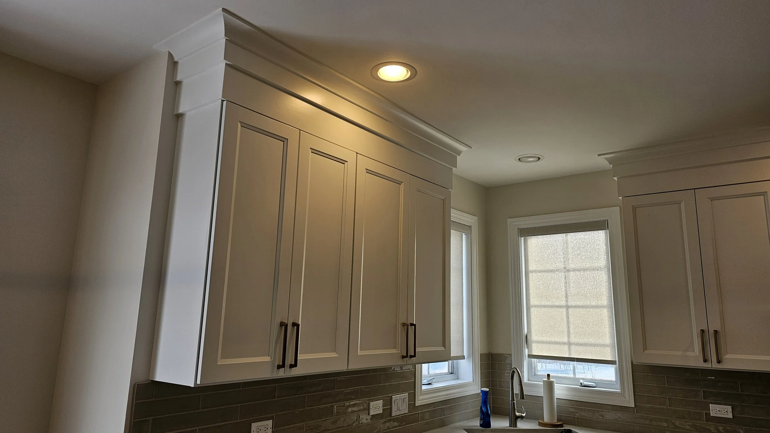Kitchen with white upper cabinets, beige walls, a window above the sink, and a faucet. There are two windows with blinds, a blue bottle, and a paper towel on the countertop.