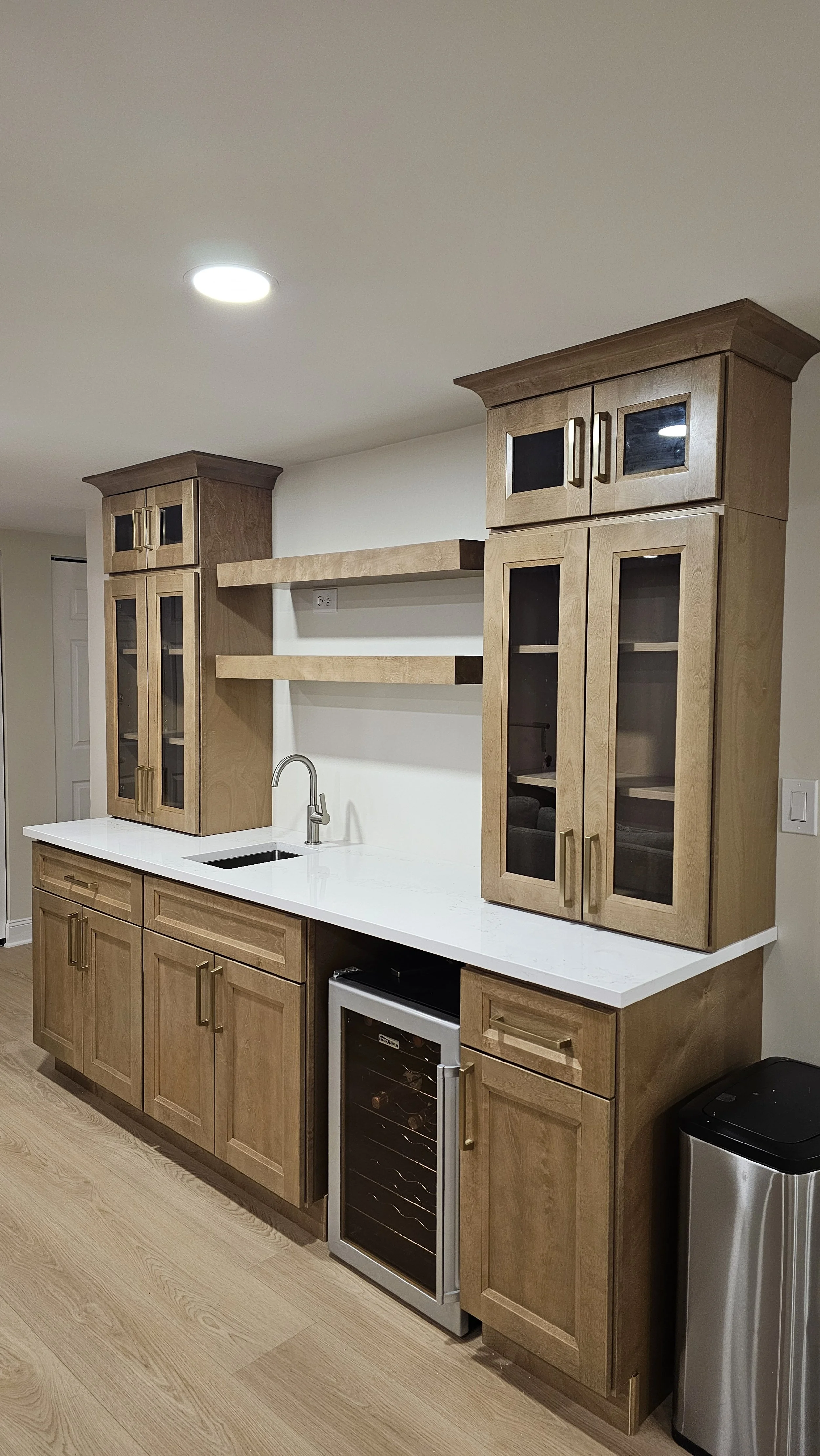 Kitchen with wooden cabinets, white countertop, small sink with stainless steel faucet, two open shelves, wine fridge, trash can, and neutral wall color.