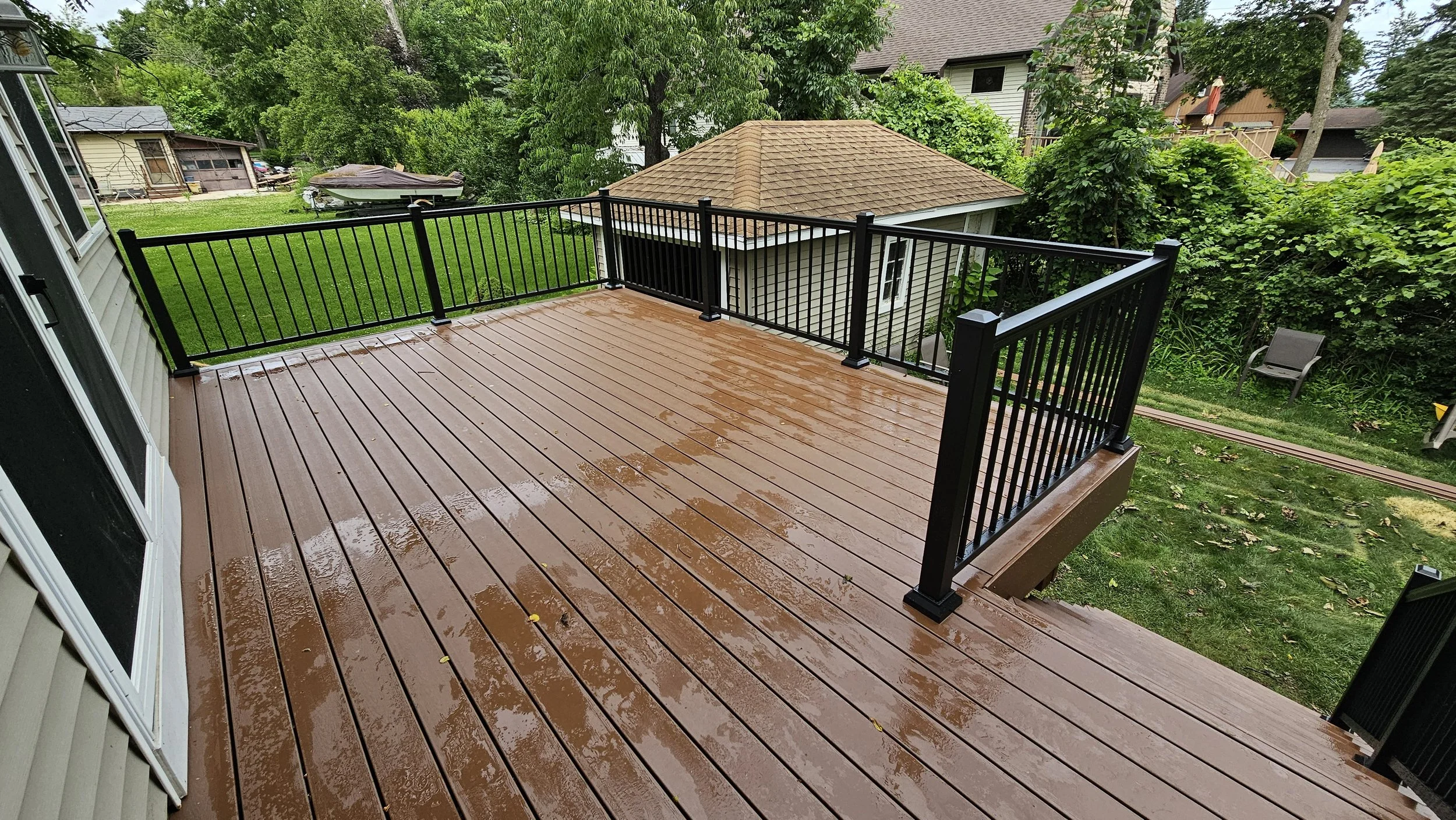 Wet wooden deck with black metal railing in a backyard with lush green trees and grass, neighboring houses, and a boat covered with a tarp.