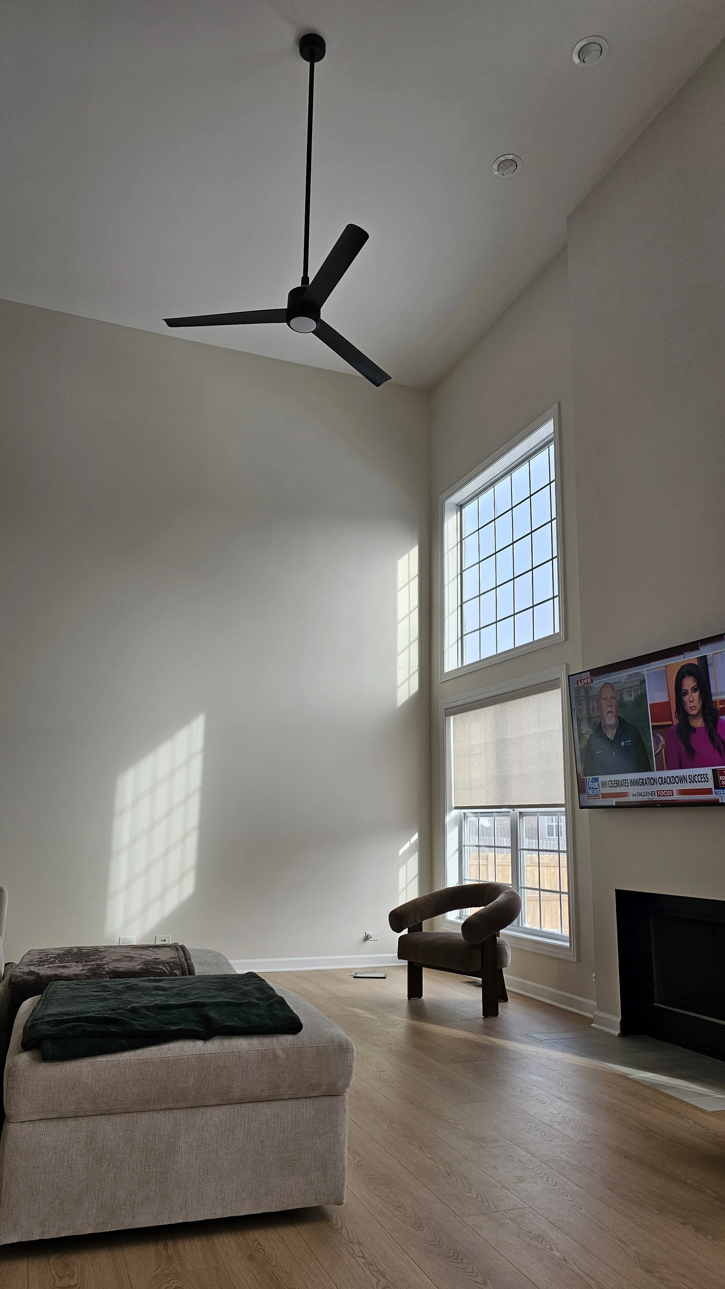 A minimalist living room with high ceilings, large windows casting sunlight patterns on the wall, a black ceiling fan, a beige ottoman with a blanket, a brown curved chair near the window, and a mounted TV showing a news broadcast.