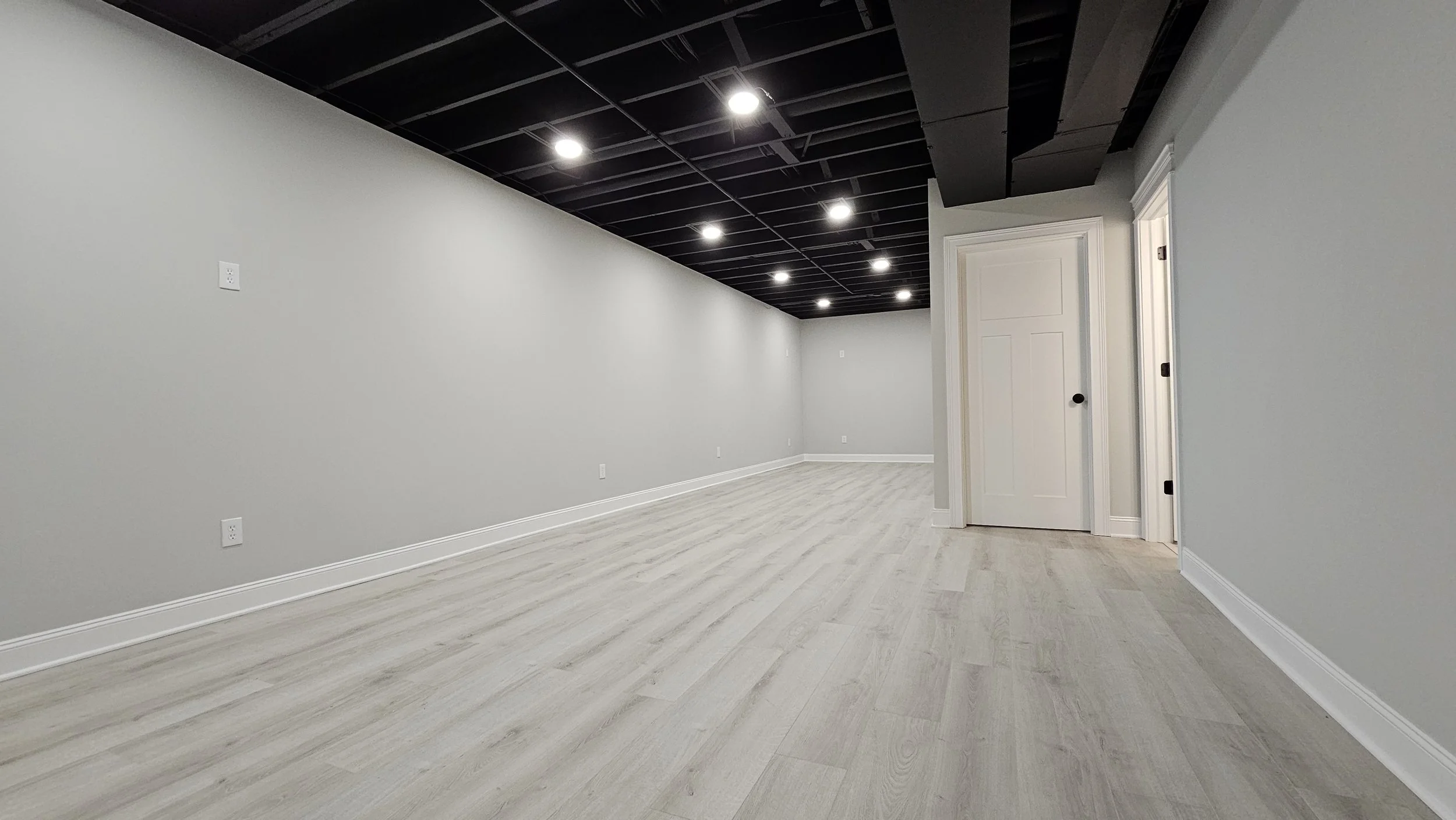 Empty finished basement with light wood flooring, grey walls, black ceiling, white baseboards, and recessed lighting.
