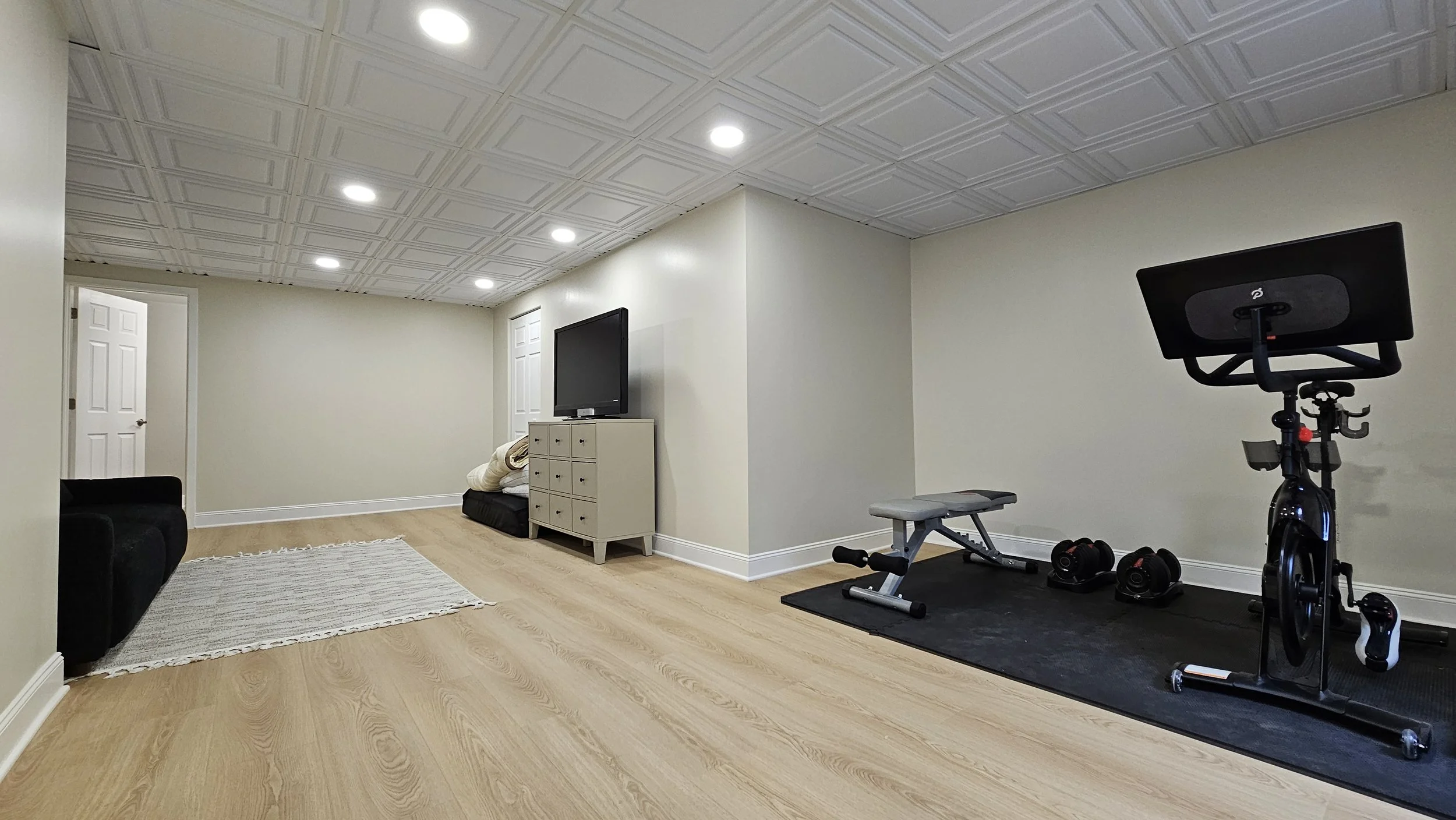 A spacious basement room with light-colored hardwood floors and white walls. The ceiling has a grid pattern with multiple recessed lights. On the right side, there is exercise equipment including a stationary bike, a treadmill, and some dumbbells on a black mat. A white cabinet with a TV on top is against the far wall. To the left, there is a small black sofa, a white/cream area rug, and an open doorway.