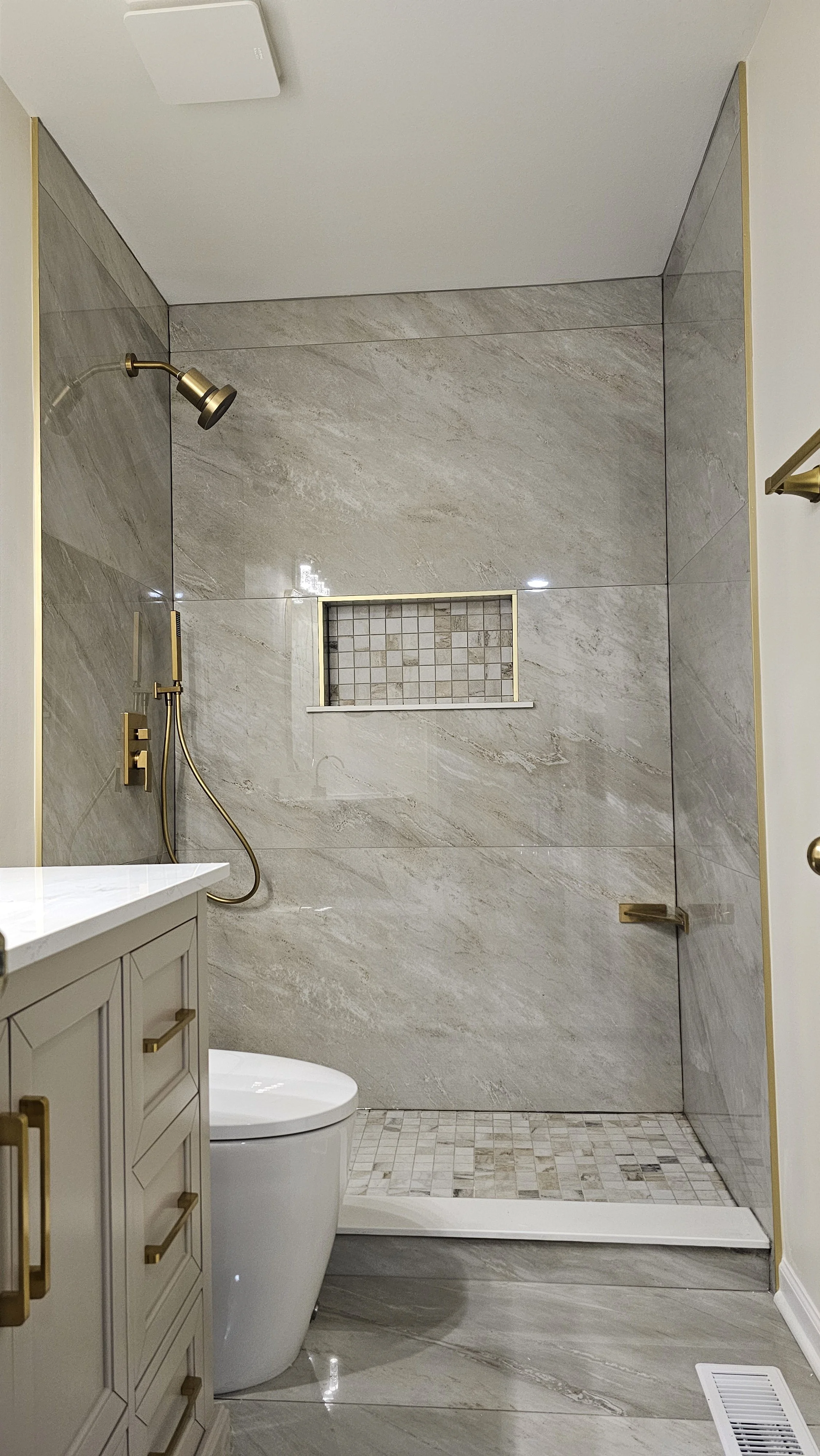 Modern bathroom with walk-in shower featuring large beige marble tiles, a small mosaic tile niche, gold fixtures, beige vanity with gold handles, and a white toilet.
