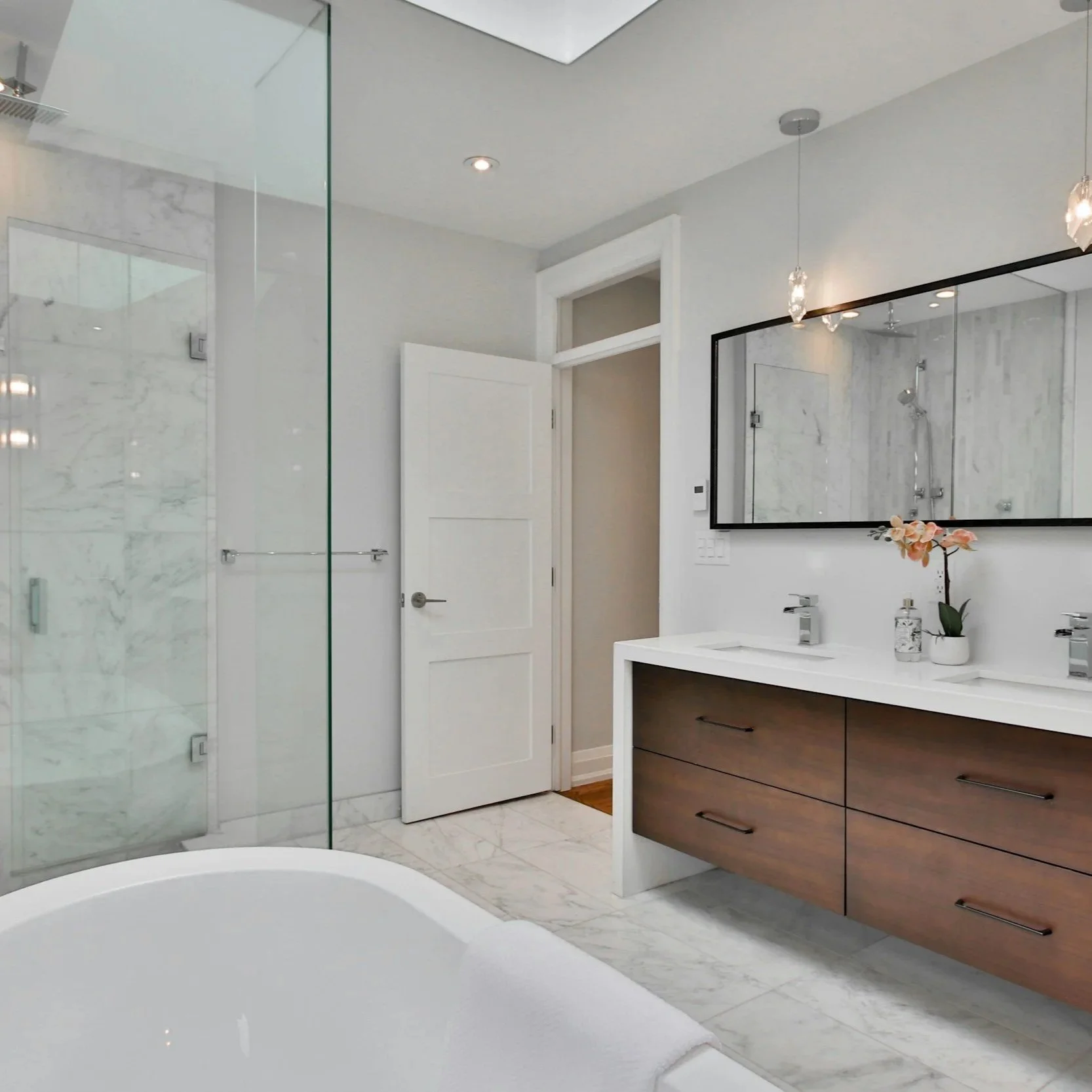 Modern bathroom with white marble flooring and walls, a glass-enclosed shower, a double vanity with a large mirror, and a bathtub with a towel.