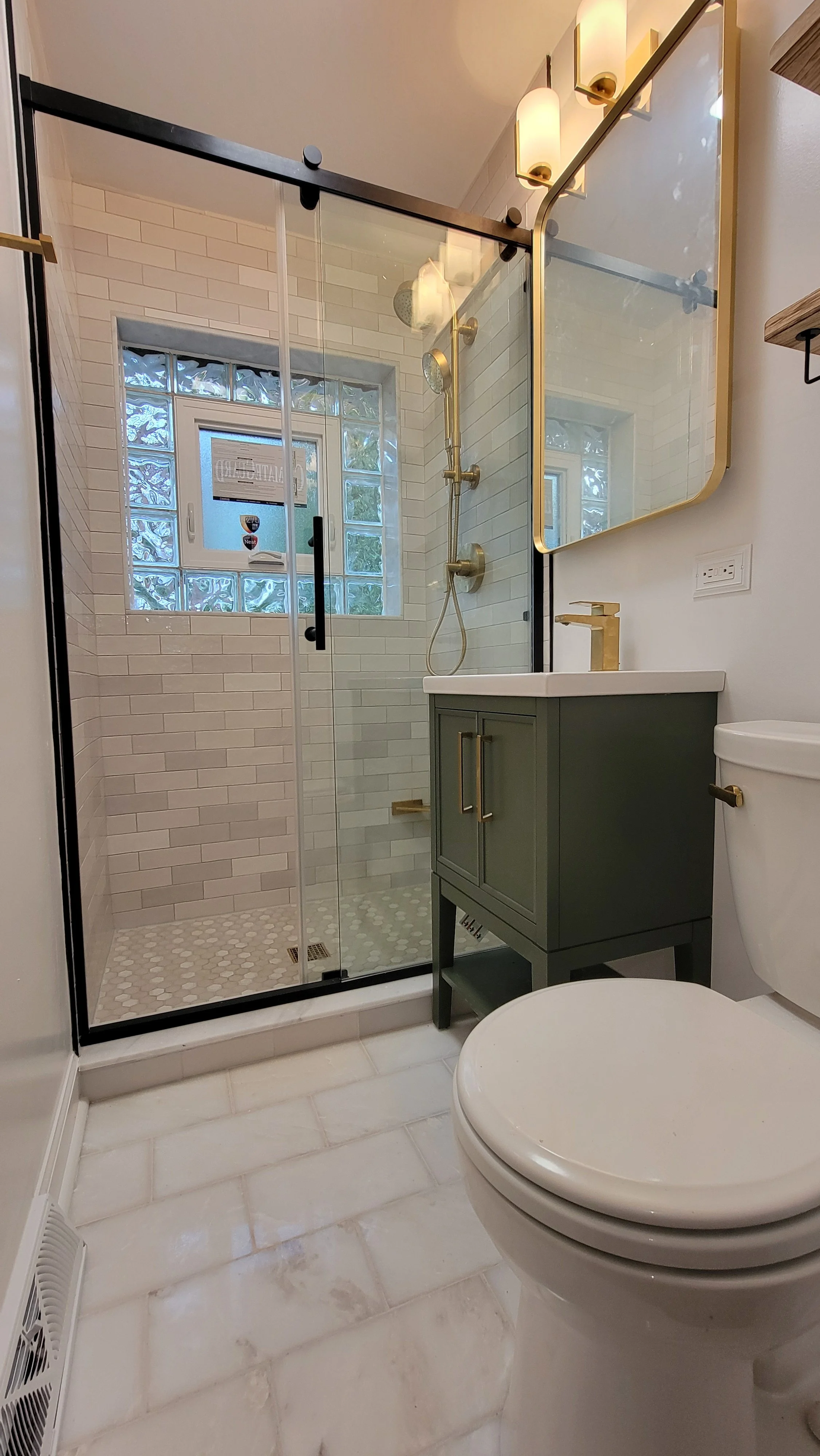 A modern bathroom with a glass-enclosed shower, a small green vanity with gold hardware, a mirror with gold trim, and a white toilet. The shower has a brass showerhead and controls, a window with frosted glass blocks, and white subway tiles. The floor is white marble-like tiles.