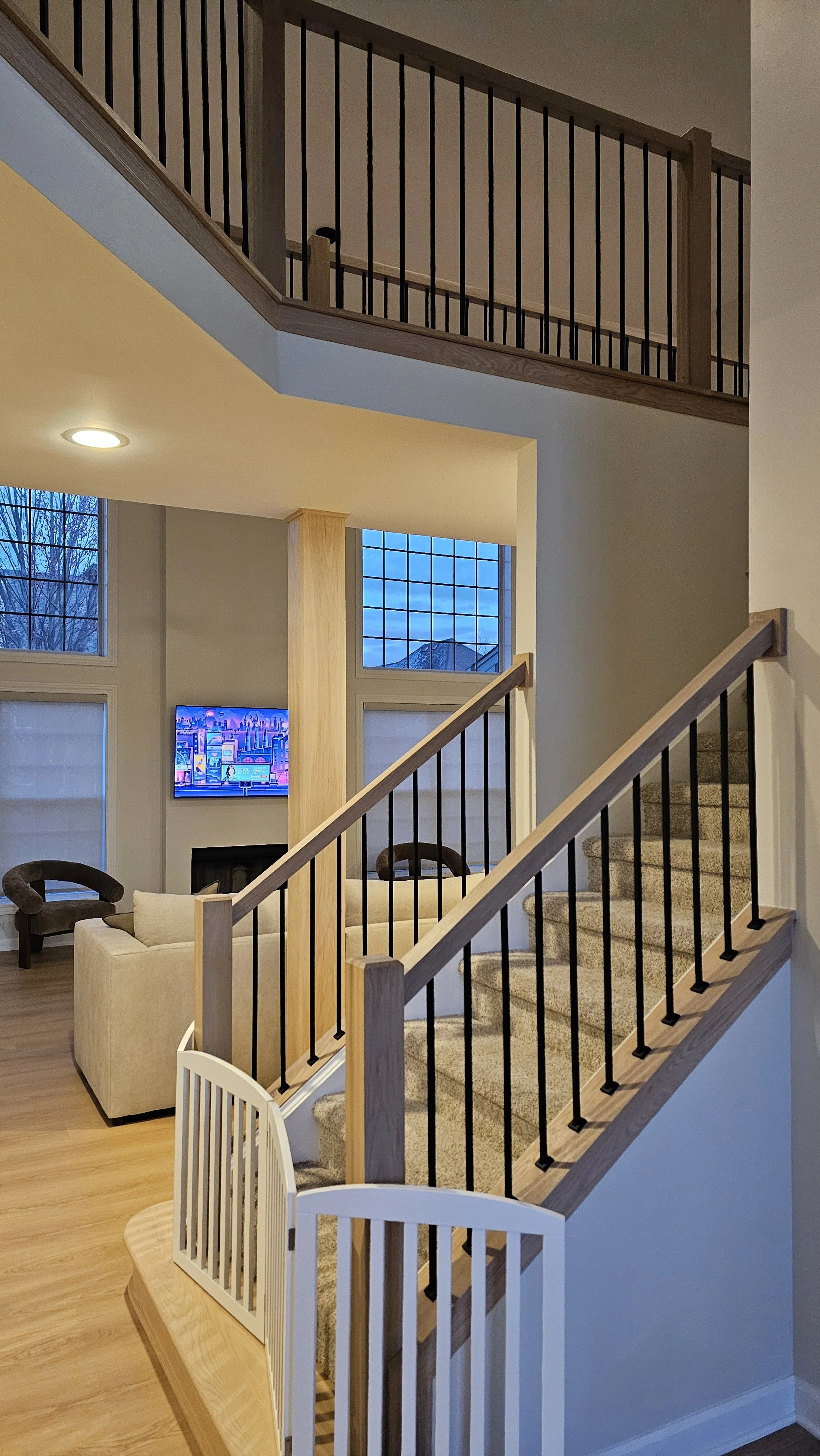 A two-story living room with wooden and black metal staircase, a beige sofa, a TV, and large windows showing a cloudy sky.