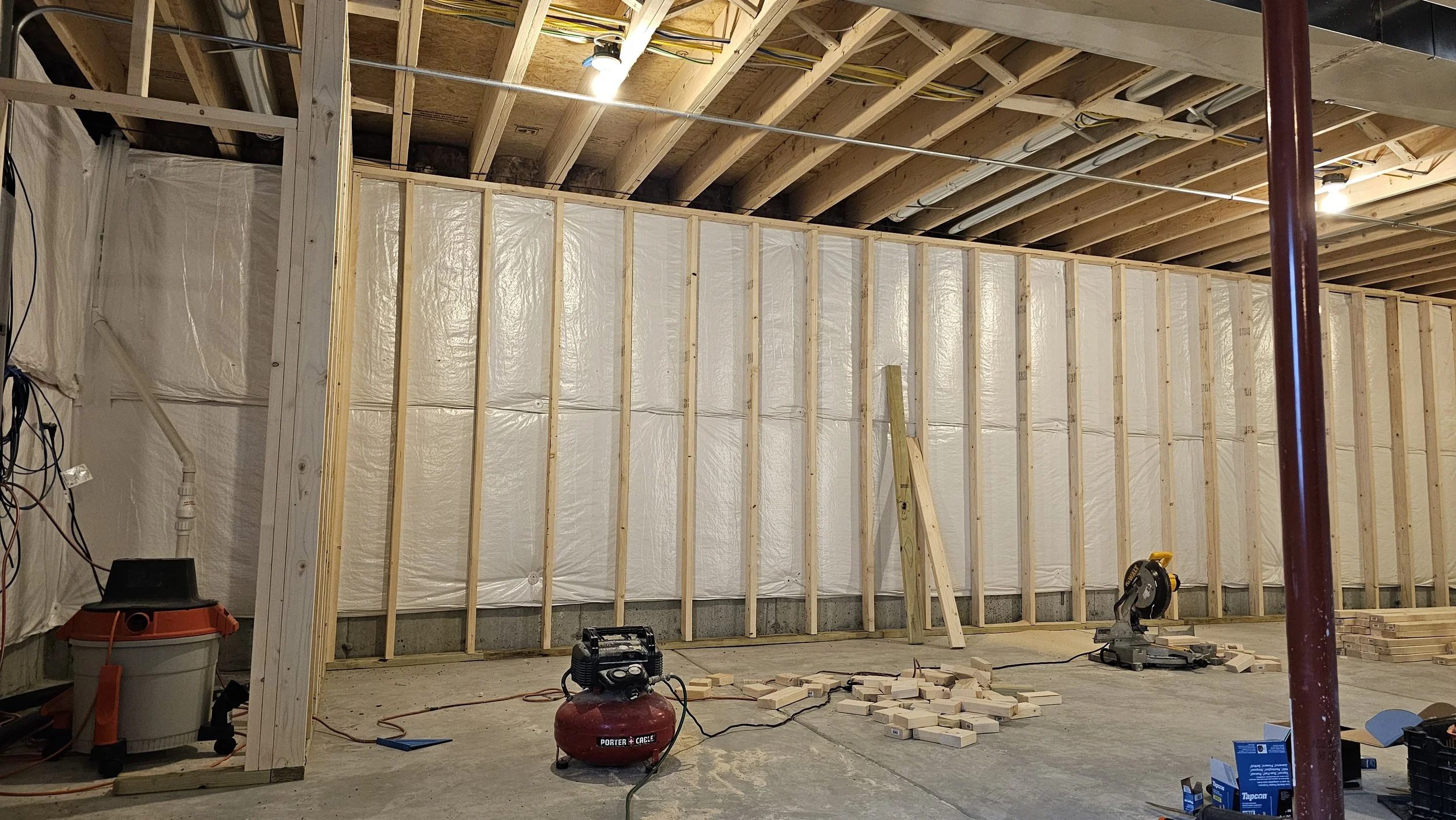 Construction site showing wooden framing, insulation, and tools, including a miter saw and air compressor, in a basement or room under renovation.