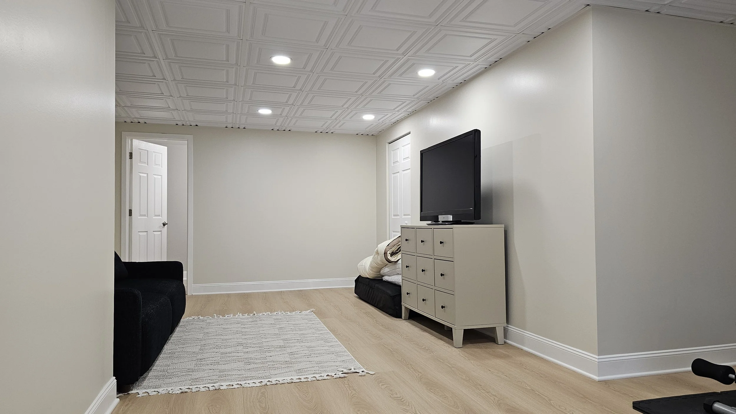 Living room with beige walls, white ceiling with recessed lights, light wood flooring, black sofa, white dresser with a television, folded blankets on a black item, a rug, and a door in the background.