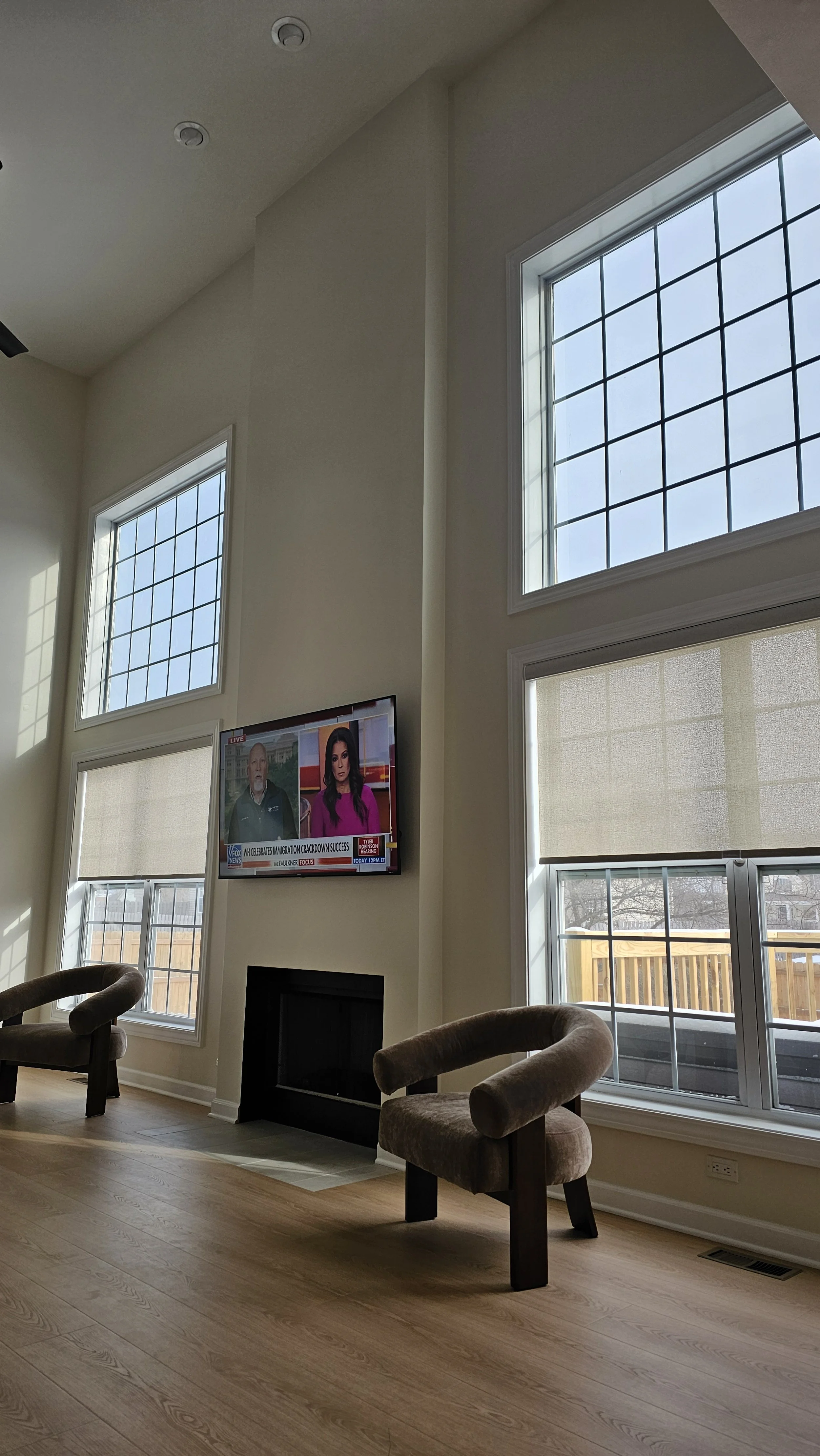 Living room with tall windows, a wall-mounted television, a fireplace, and two brown upholstered chairs, with a view of a backyard.