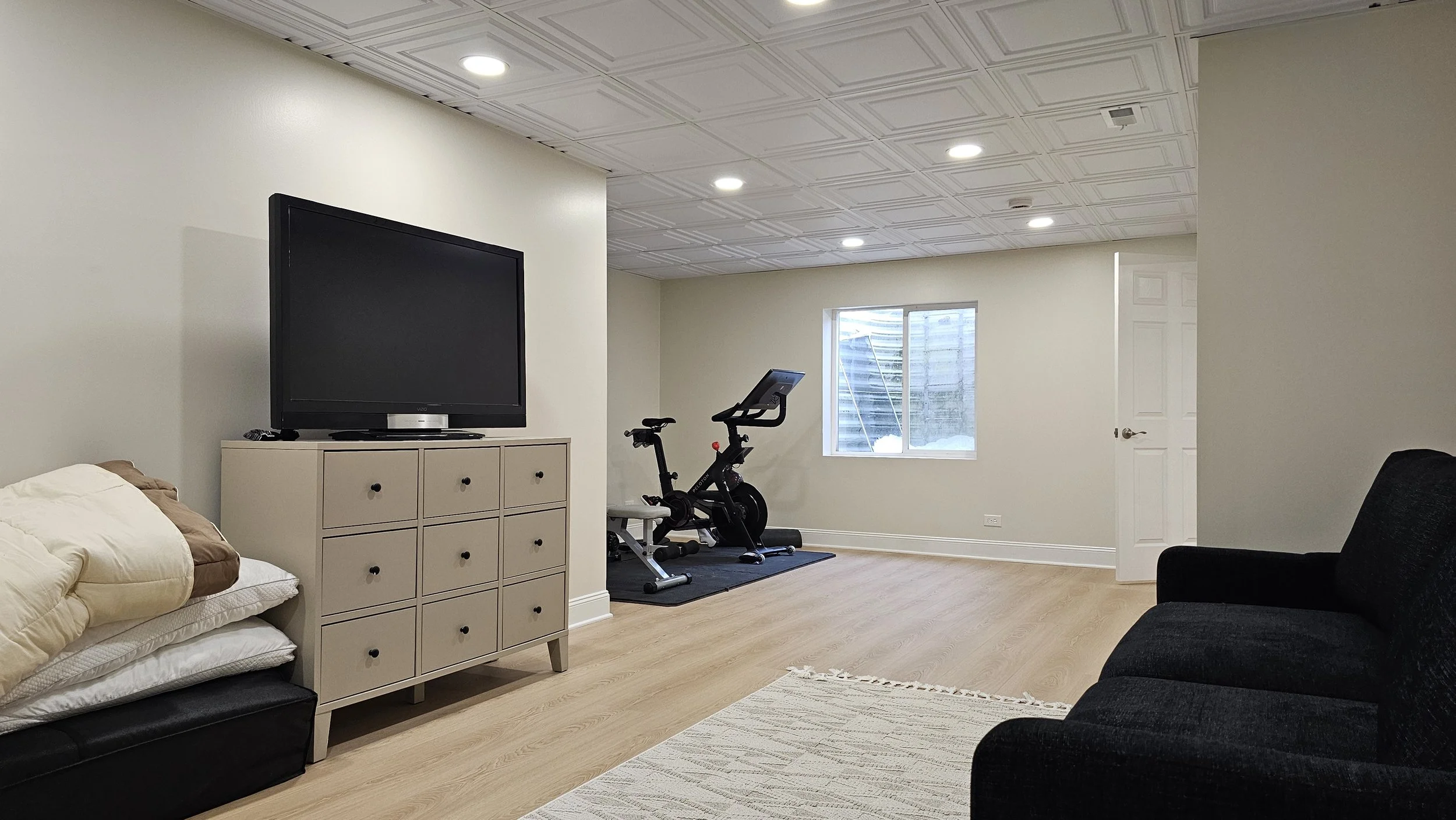 Living room with beige walls, wooden floor, a large flat-screen TV on a beige cabinet, a black exercise bike on a black mat, a window with a snowy outdoor scene, a closed door, a black sofa, a beige rug, and ceiling lights.