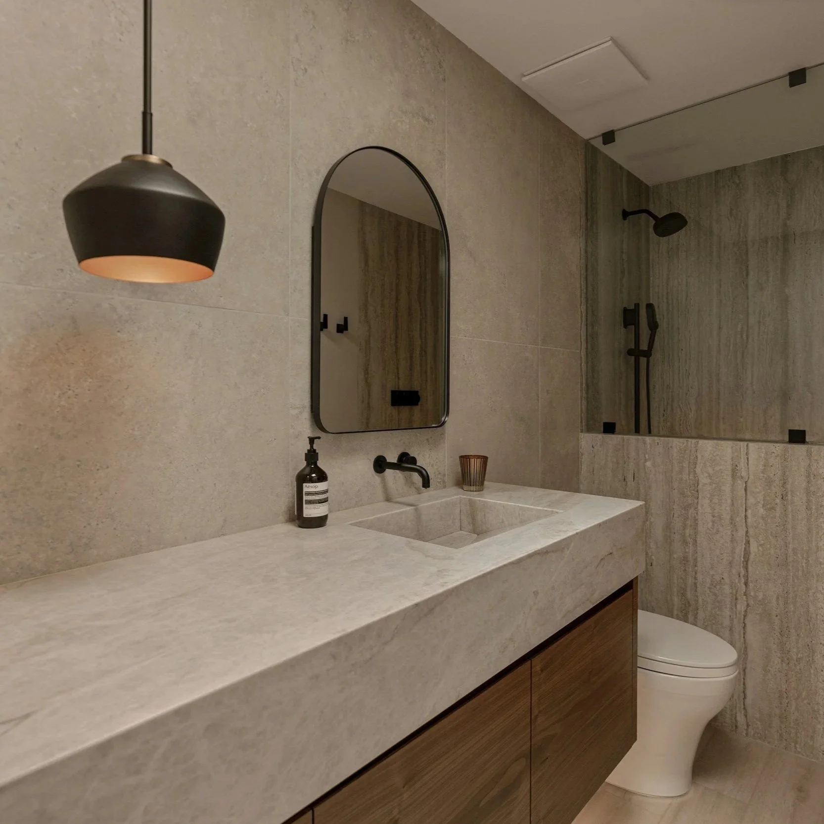 Modern bathroom with a beige stone countertop, black fixtures, and a shower area with a rain shower head. Mirror above the sink, soap dispenser, and a small container on the countertop. Toilet to the right of the sink.