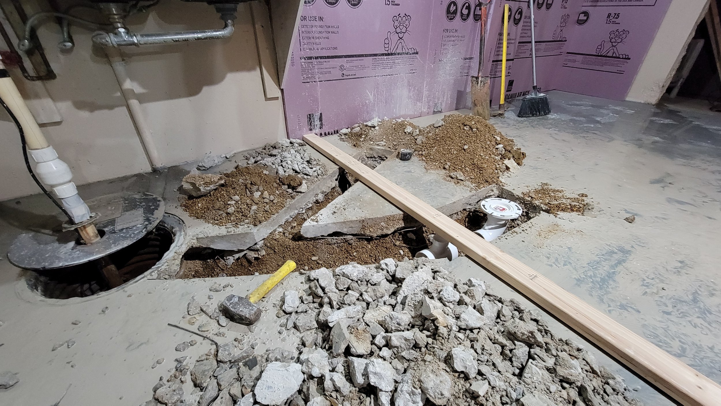 Home bathroom under construction with exposed pipes, open floor with debris, gravel, APEX plumbing fixture, wooden board across the floor, pink insulation panels on the wall, and cleaning tools in the background.