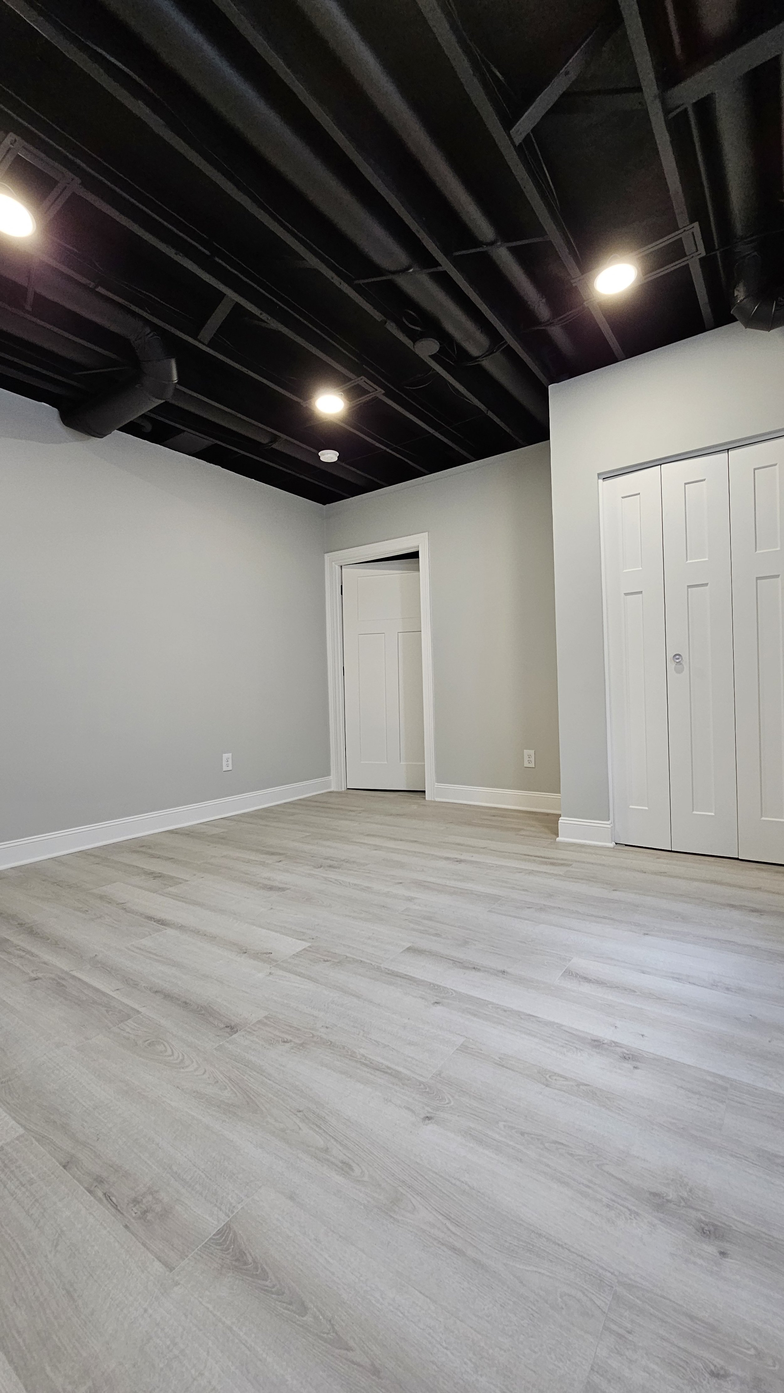 Empty room with light-colored hardwood floors, light gray walls, white baseboards, a white door, and a closet with white bifold doors. The ceiling is black with visible ducts and recessed lighting.