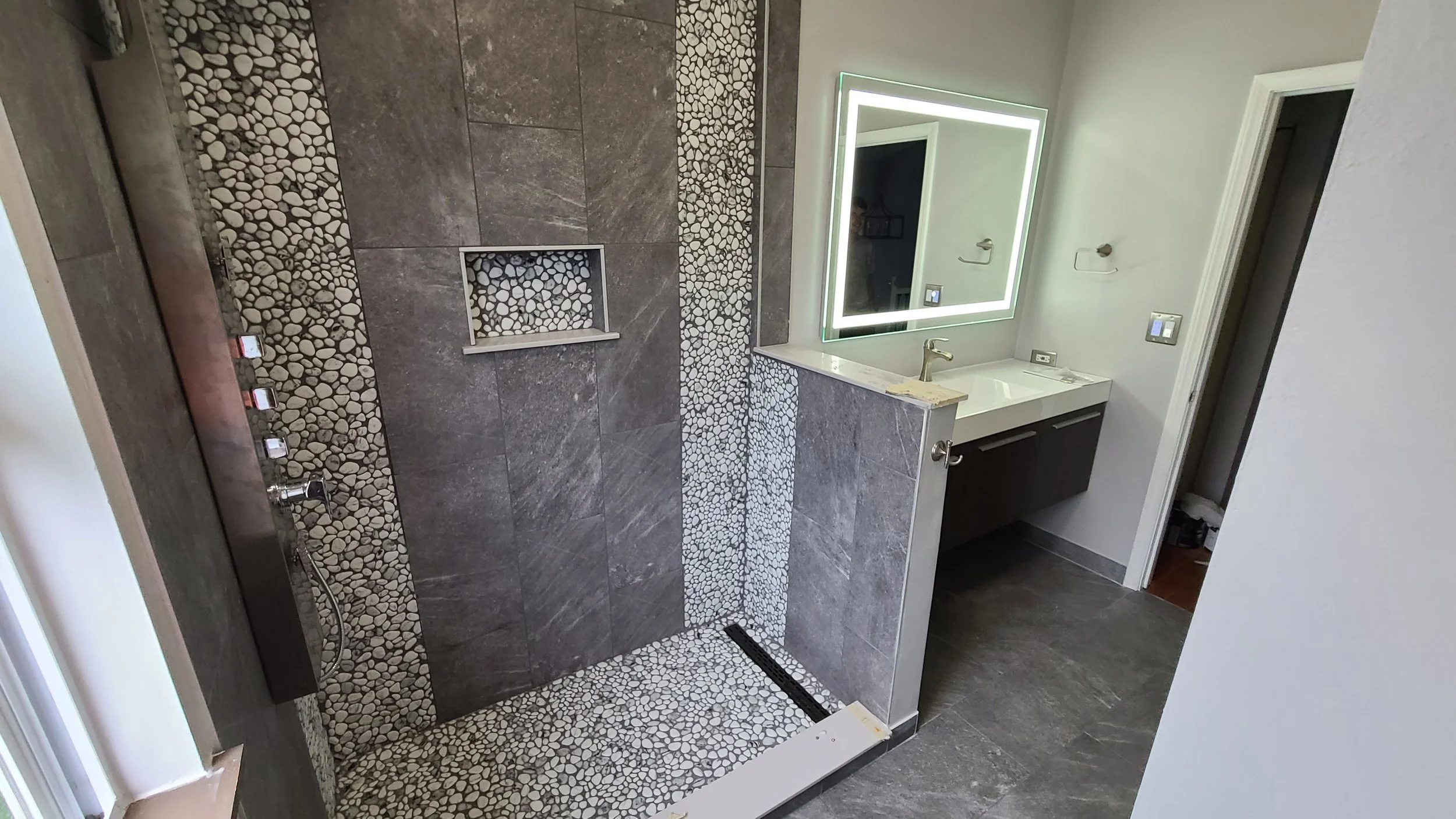 Modern bathroom with pebble stone and gray tile shower, illuminated mirror above white sink, dark vanity, and open door leading to another room.