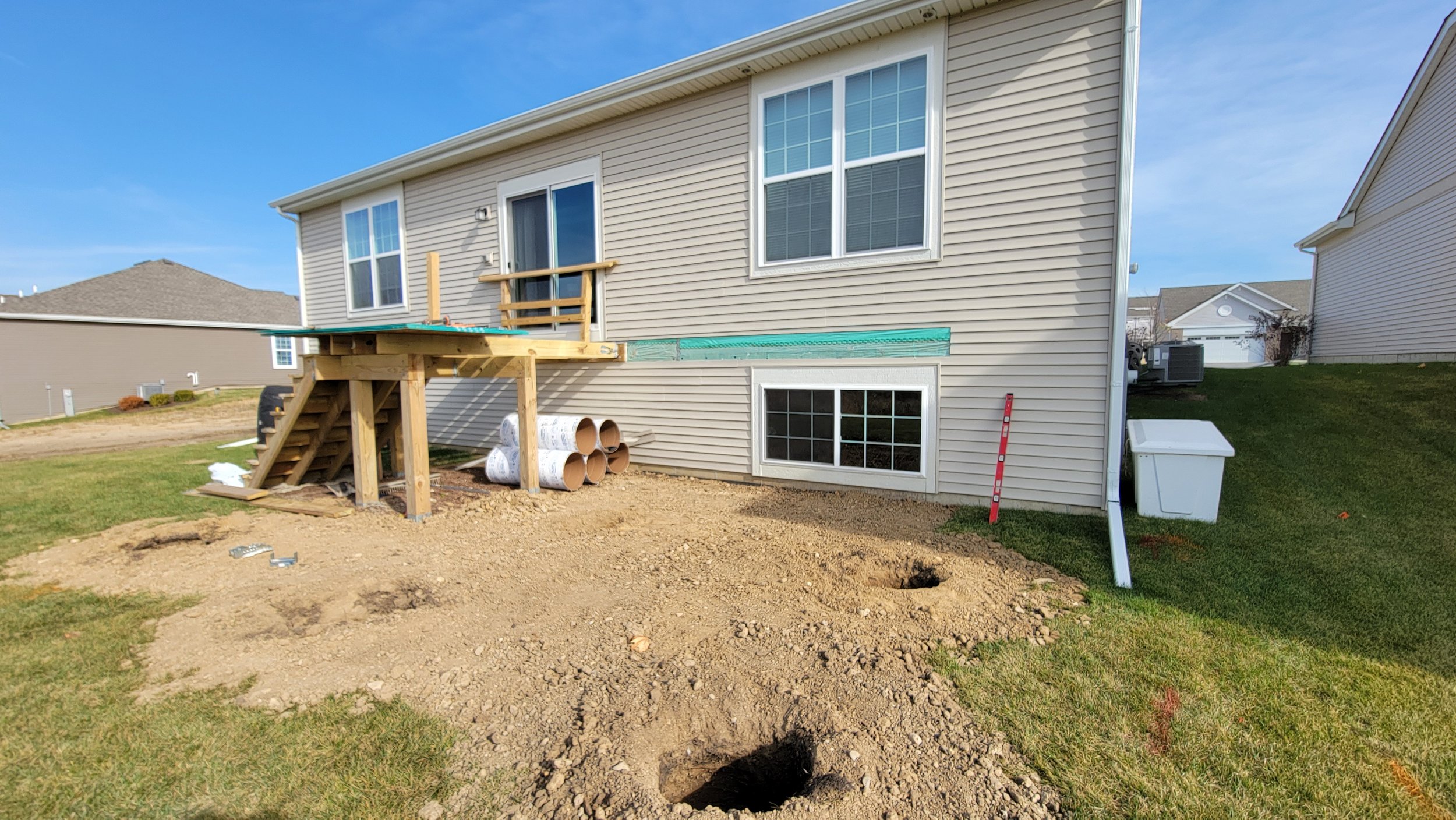 Backyard of a house under construction with a new wooden deck being built, dirt and holes in the ground, construction materials, and neighboring houses visible.