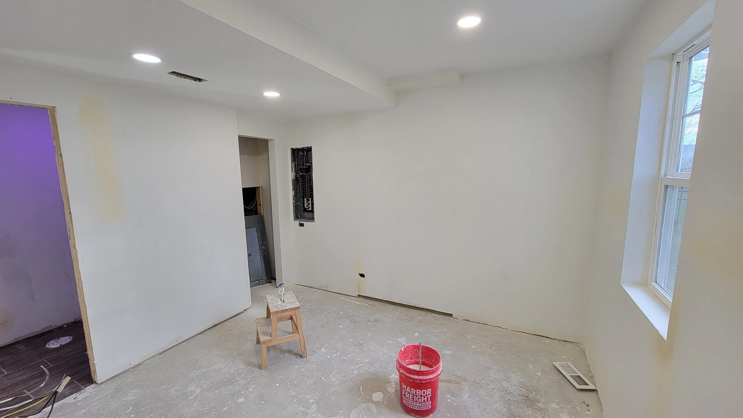 Interior of a room under renovation with white walls, a window on the right side, and construction materials on the floor including a small wooden stool, a red bucket, and electrical wiring.