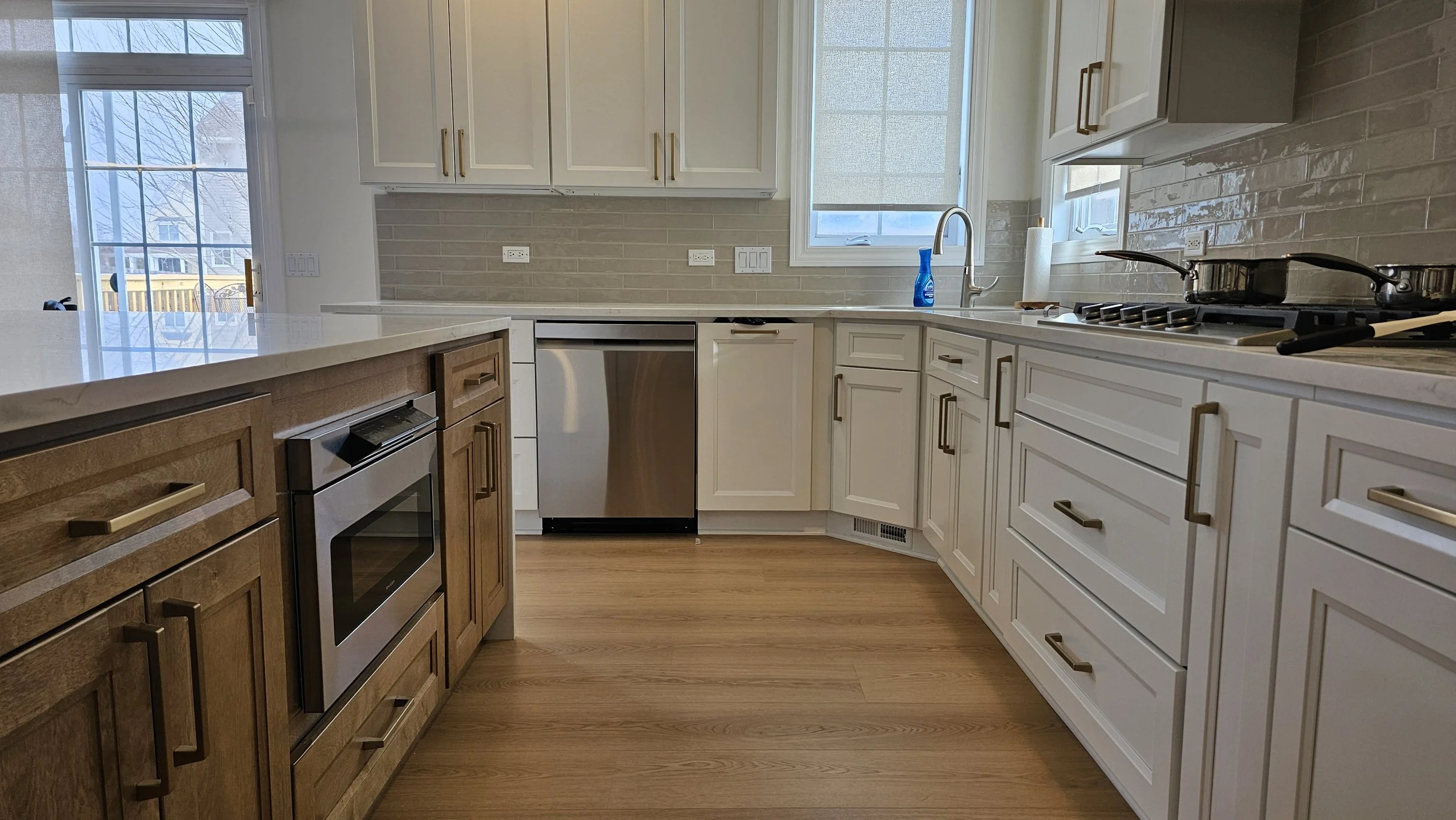 Modern kitchen with white and wood cabinets, stainless steel oven and dishwasher, gray countertops, and light-colored hardwood floors.