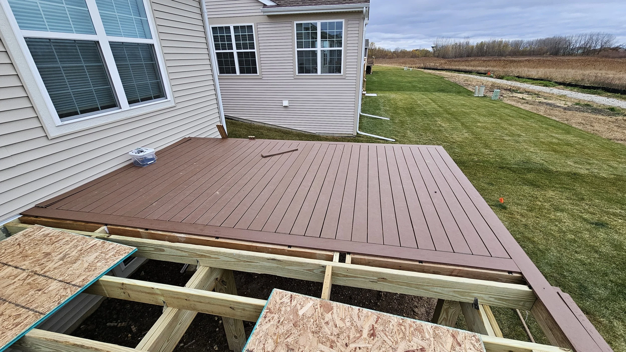 View of a partially built wooden deck with brown composite decking boards attached to the house, with grassy backyard and a pathway in the background.