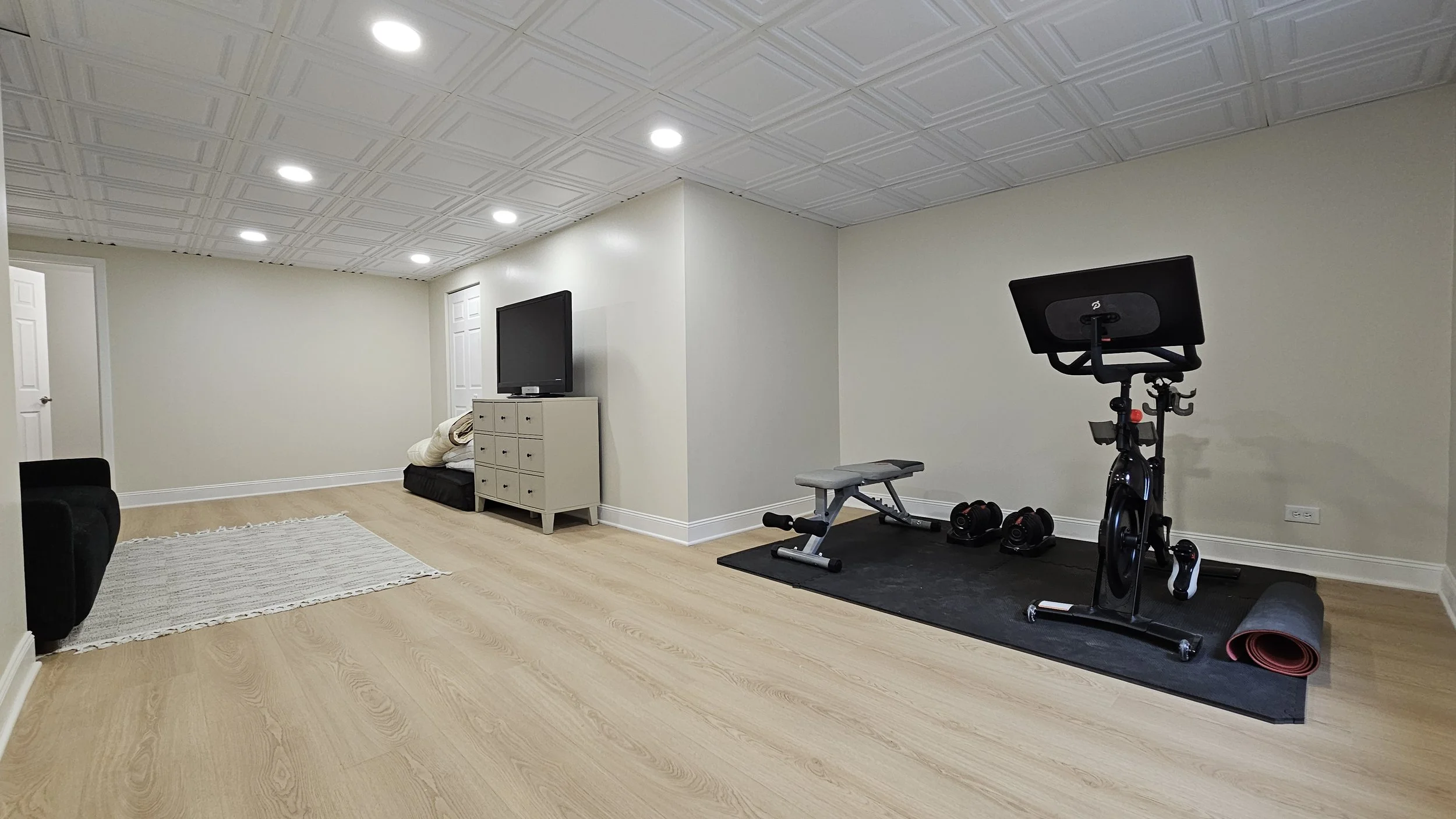 Empty basement room with wood laminate flooring, white walls, and a white ceiling with recessed lighting. There is a black armchair on the left, a white dresser with a TV on top in the center, and a small black and gray exercise area with a stationar