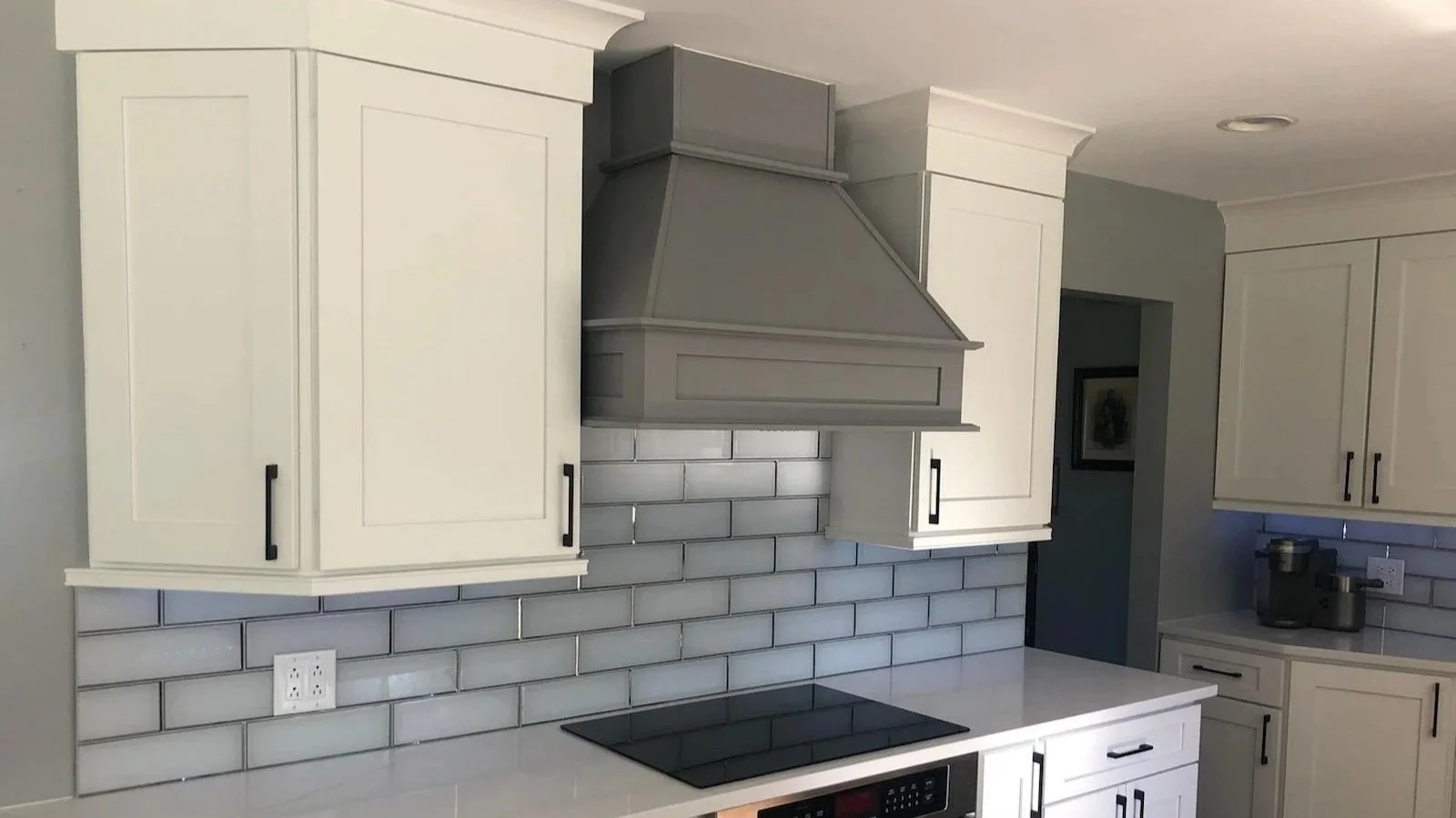 Modern kitchen with white cabinets, gray range hood, gray subway tile backsplash, and black handles, with an electric cooktop on the white countertop.