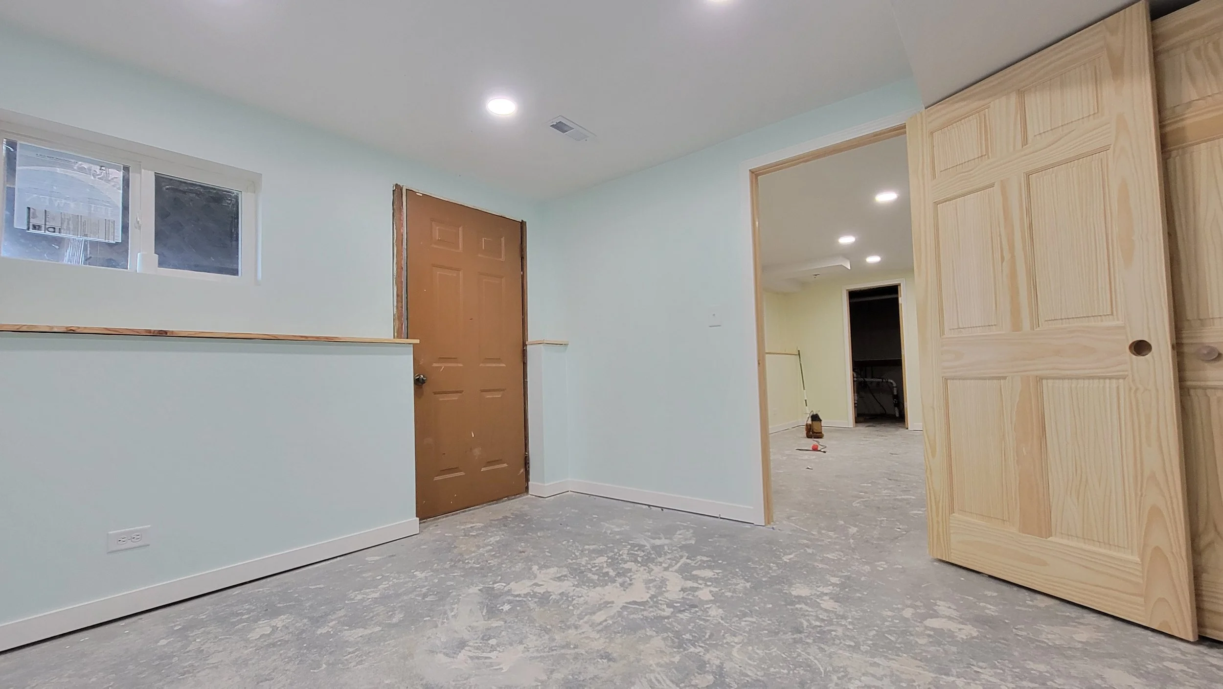 Empty room under construction with a small window, a brown door, and an unfinished light wood closet door. The floor is concrete with construction dust.