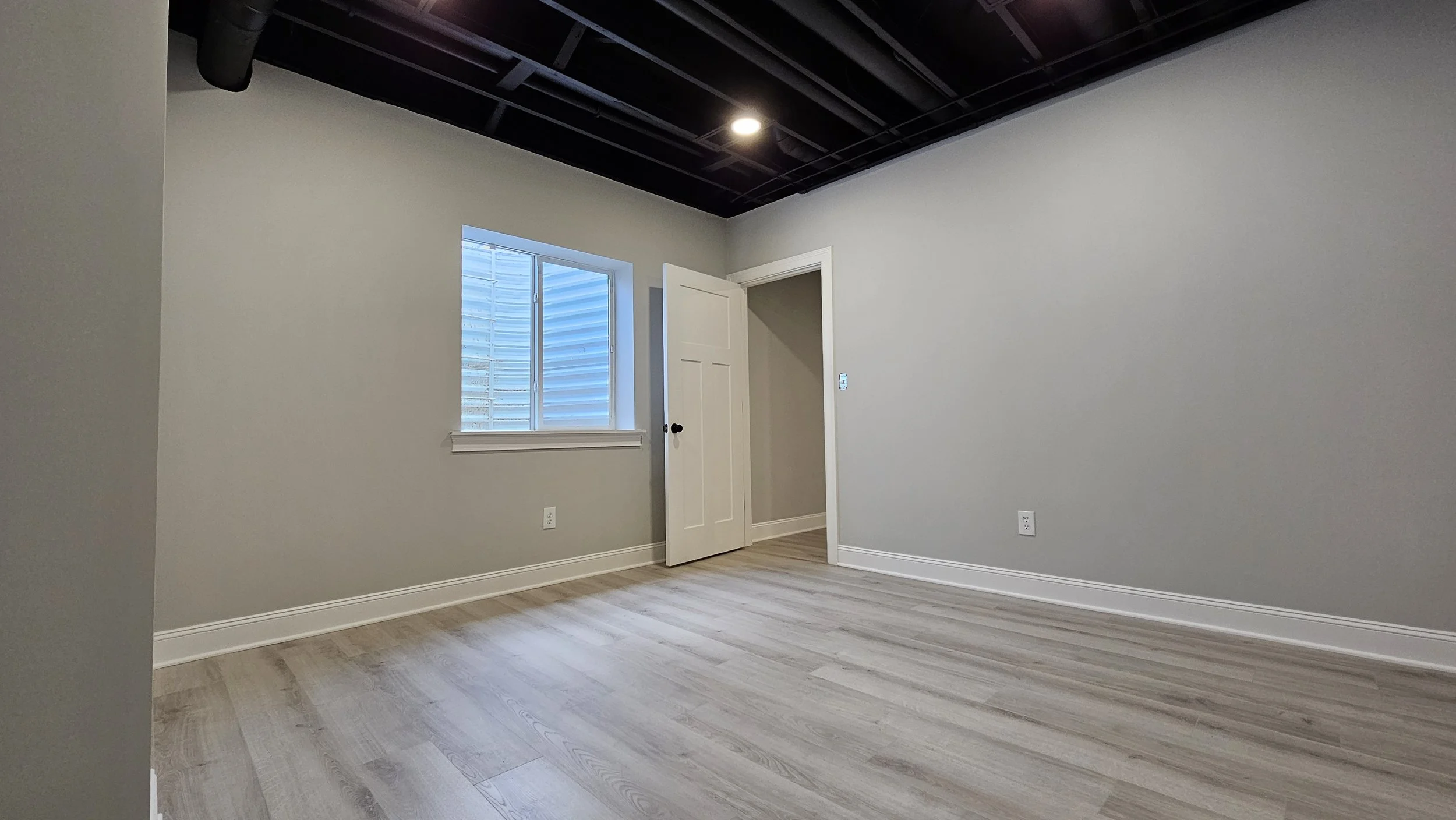 Empty room with light-colored wooden flooring, gray walls, a window with blinds, an open door leading to a small closet, and a ceiling with black grid pattern and ceiling light.
