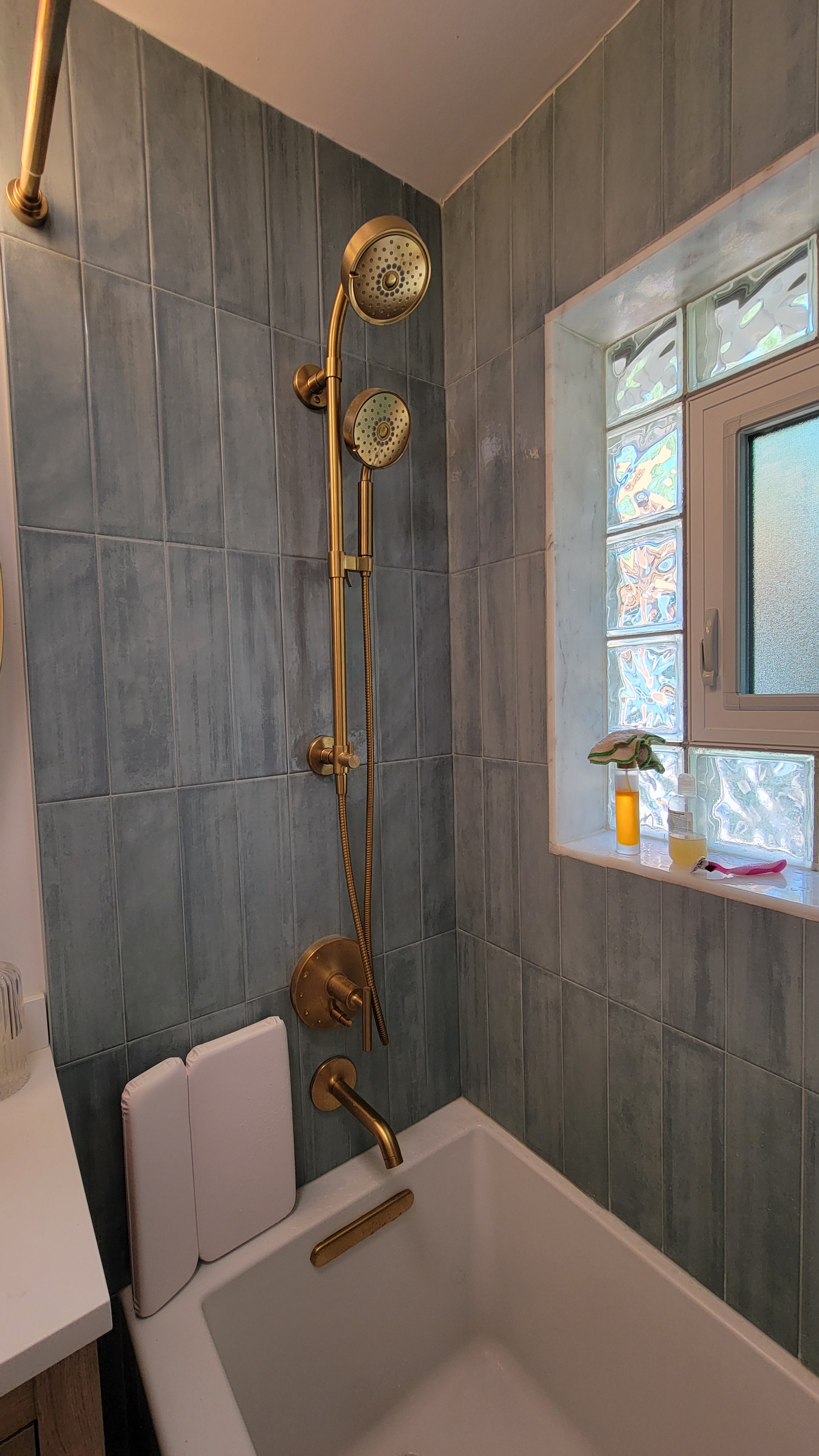 A bathroom shower with green tiles, a brass showerhead and fixtures, a small window with glass blocks, and various bottles and items on the windowsill.
