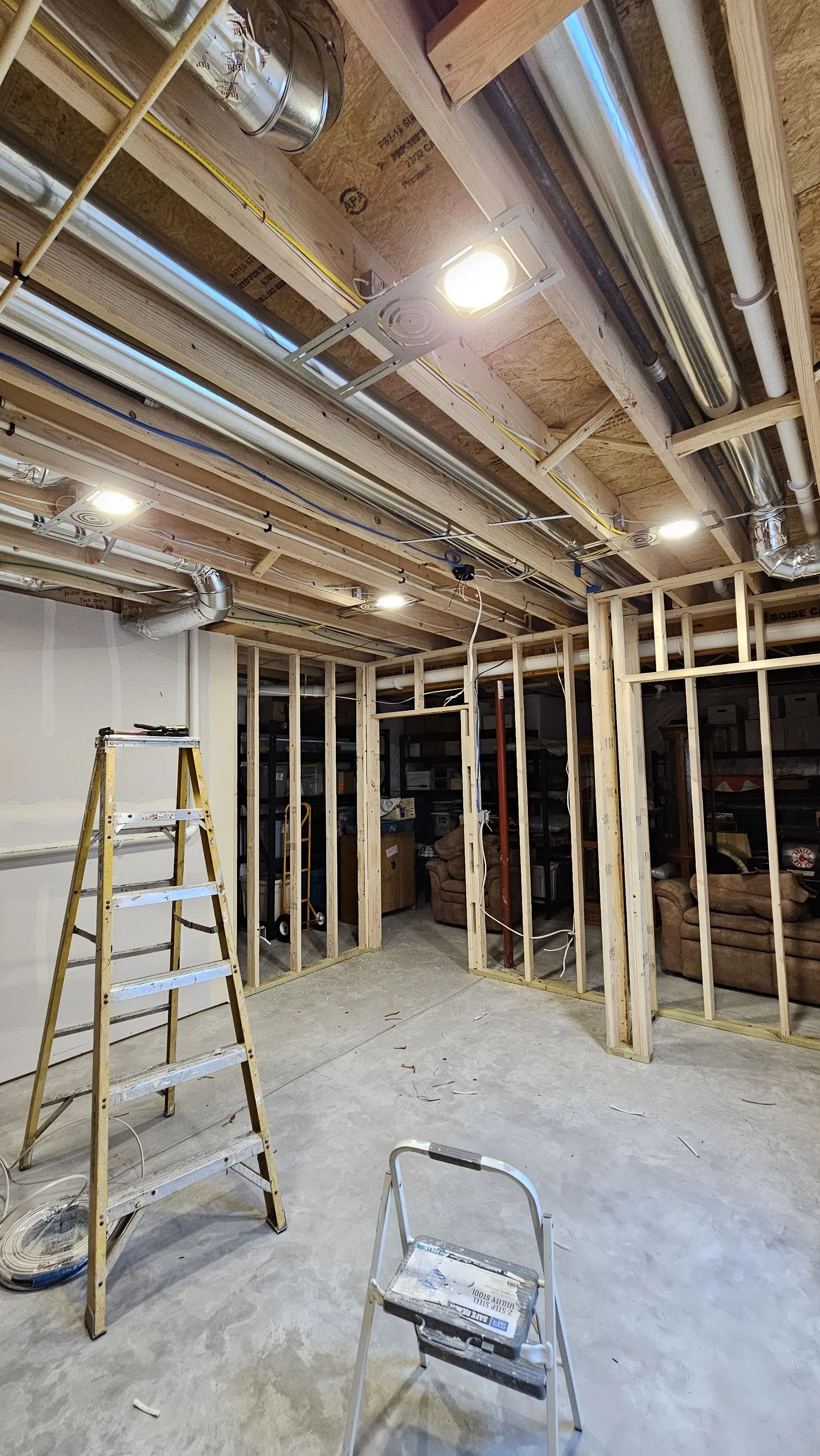A basement under construction with unfinished walls and ceiling framing. There are two ladders, ceiling lights, and exposed pipes and electrical wiring visible.