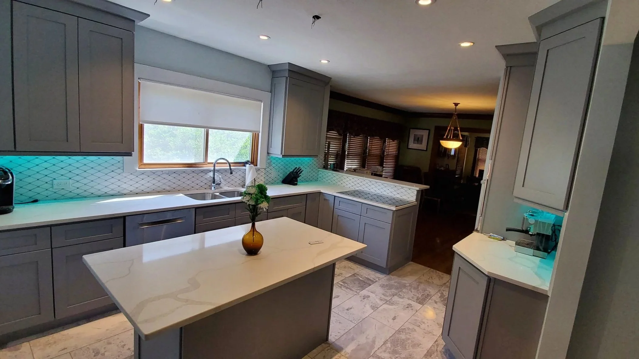 Modern kitchen with gray cabinets, white countertops, and a tile backsplash. A window above the sink lets in natural light. There is a small kitchen island with a vase of flowers. The floor is tiled. In the background, a dining area with wooden blinds and a chandelier is visible.