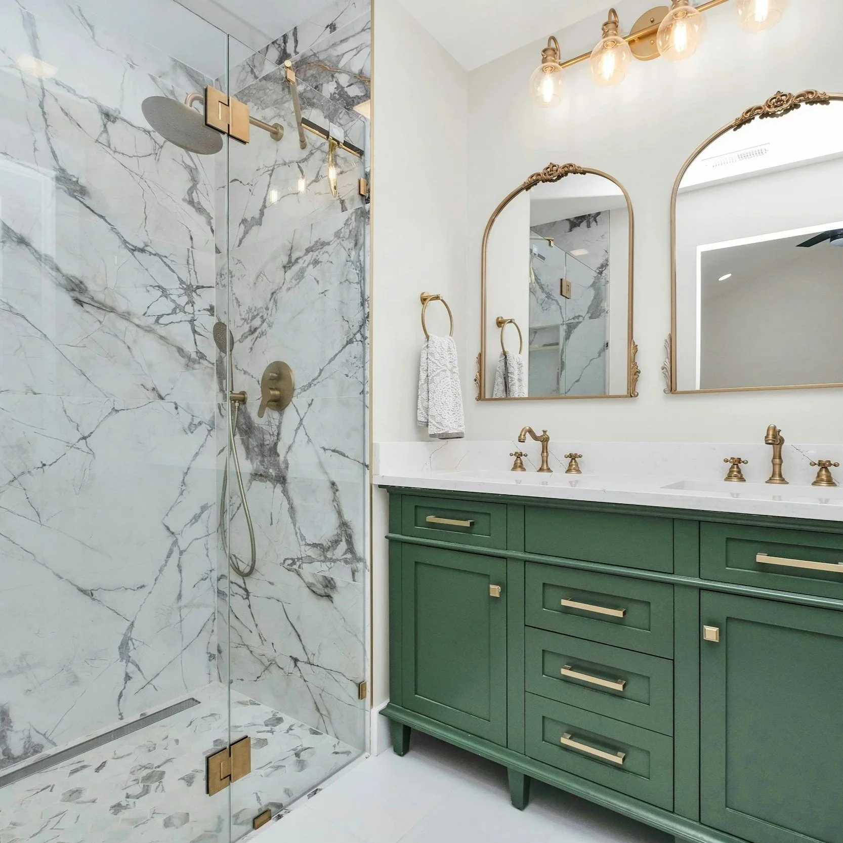A modern bathroom with a marble shower enclosure, green vanity with gold handles, and gold fixtures. Two mirrors with decorative gold frames are above the vanity, and a row of wall-mounted light bulbs is above the mirrors. A white towel hangs on a gold towel ring next to the shower.