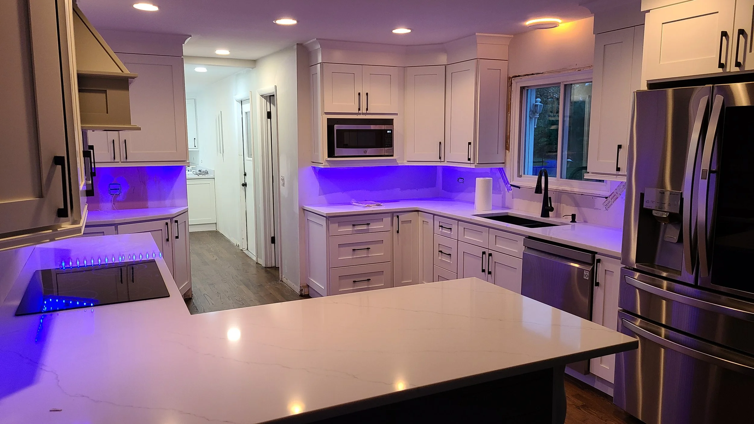 Kitchen with white cabinets, black handles, stainless steel refrigerator, microwave, and dishwasher. Purple LED lighting beneath cabinets and on stovetop. Includes window over sink with black faucet, island with white countertop, and wood flooring.