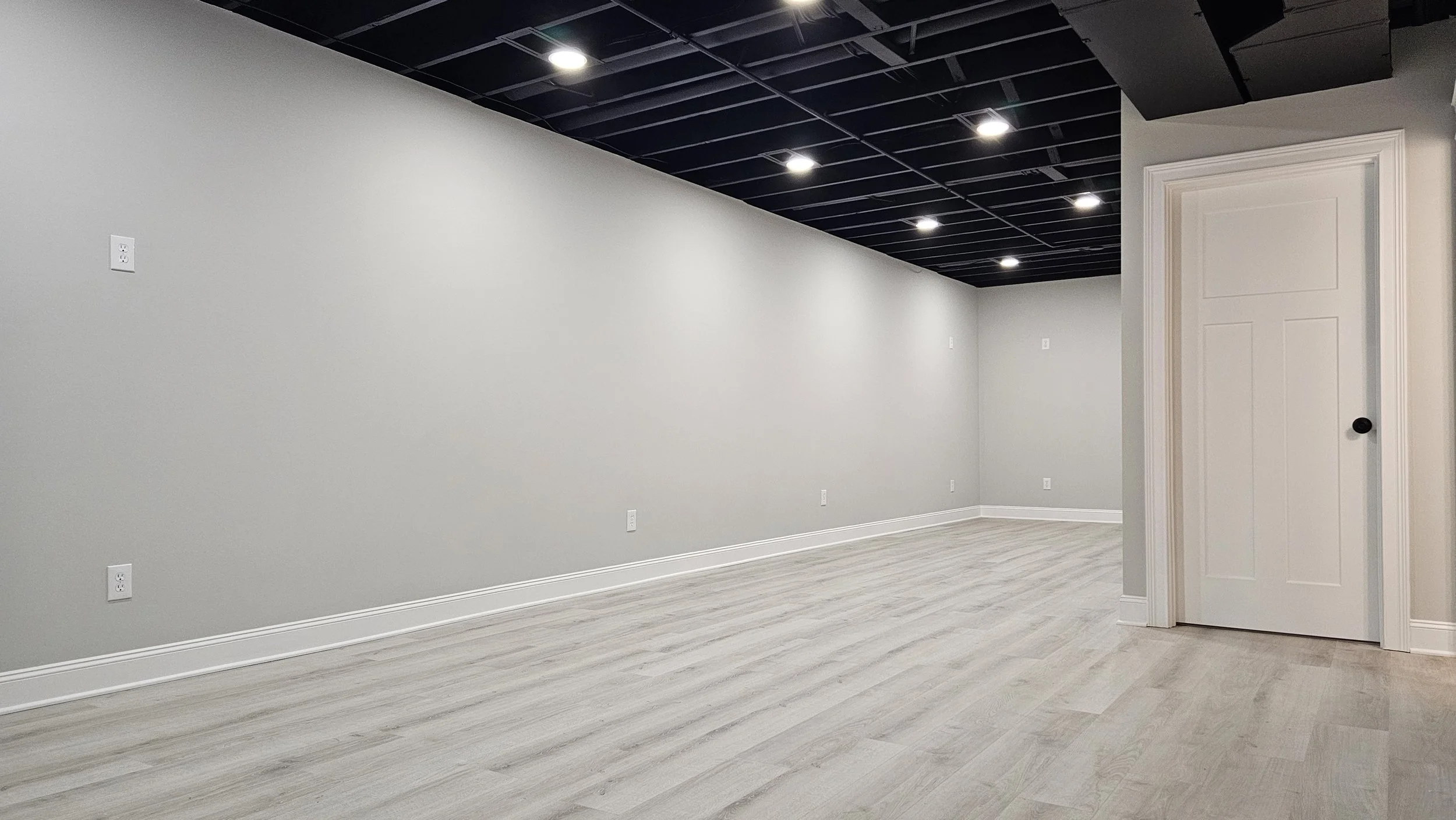 Empty room with light gray walls, light wood flooring, white baseboards, a ceiling with exposed black beams and recessed lighting, and a closed white door with molding on the right side.