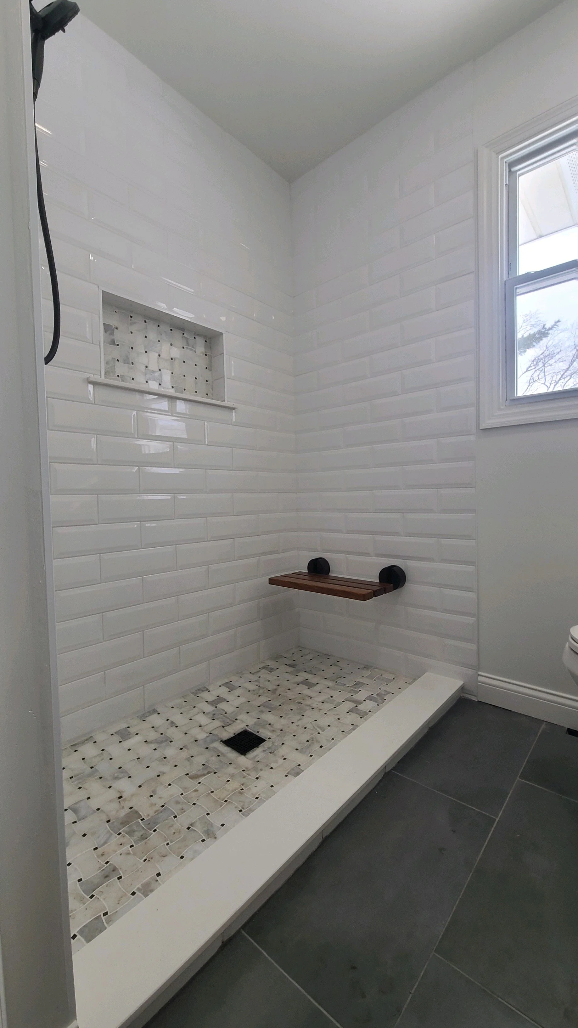 Unfinished walk-in shower with white subway tile walls, a small built-in niche, a wooden bench, and a window with a white frame