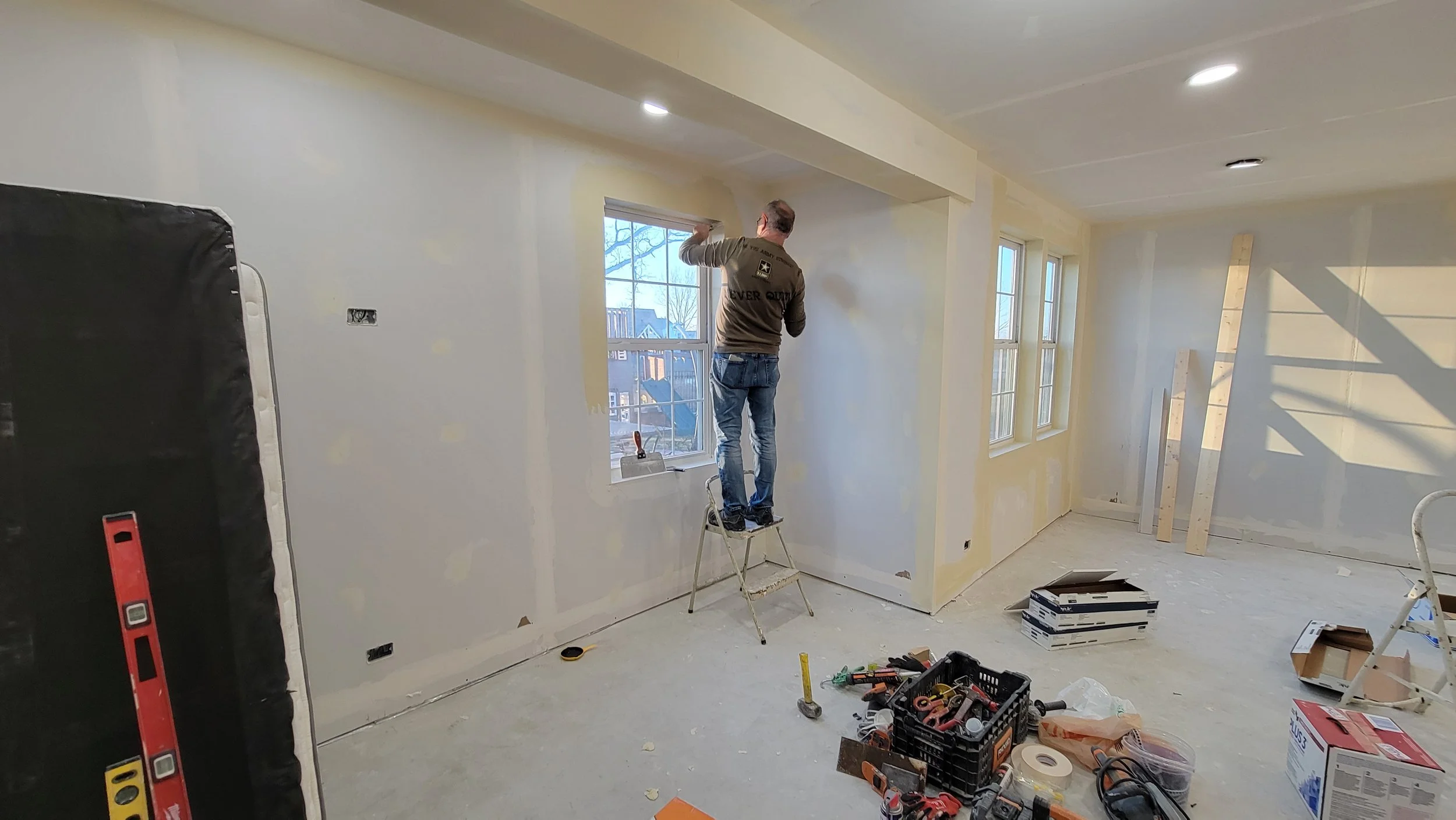 Man standing on a step ladder installing a window in a room under renovation with drywall and construction tools on the floor.