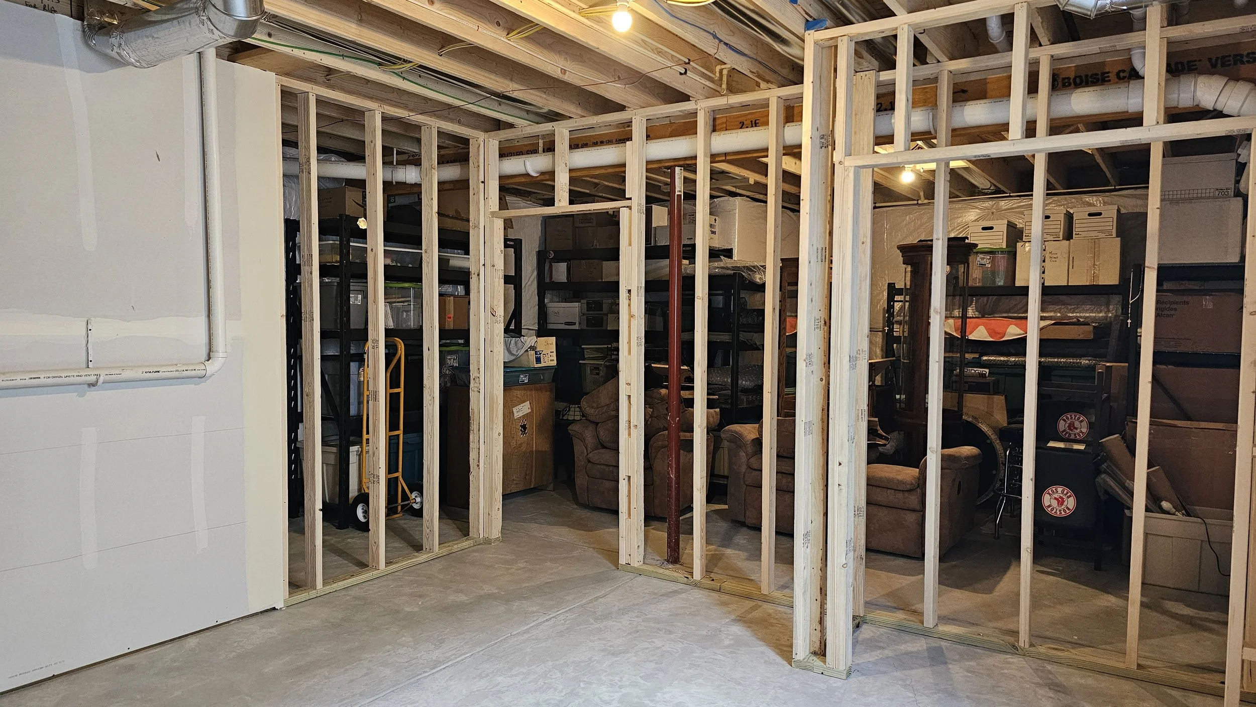Partially constructed indoor wall with wooden studs in a basement, with stored boxes, furniture, and shelving in the background.