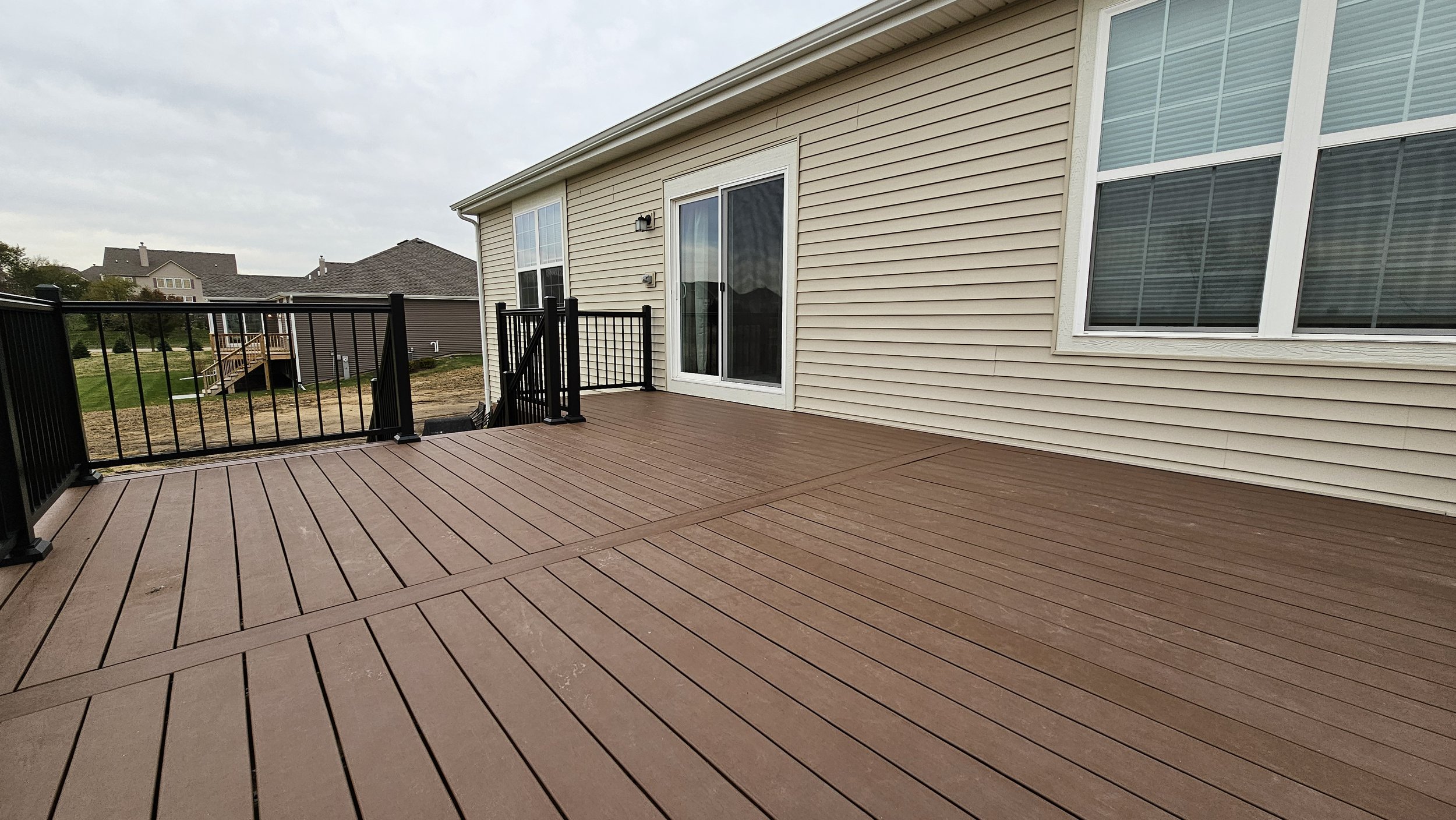 A large wooden deck attached to a house with a sliding glass door and windows, surrounded by a black metal railing, with neighboring houses visible in the background under a cloudy sky.