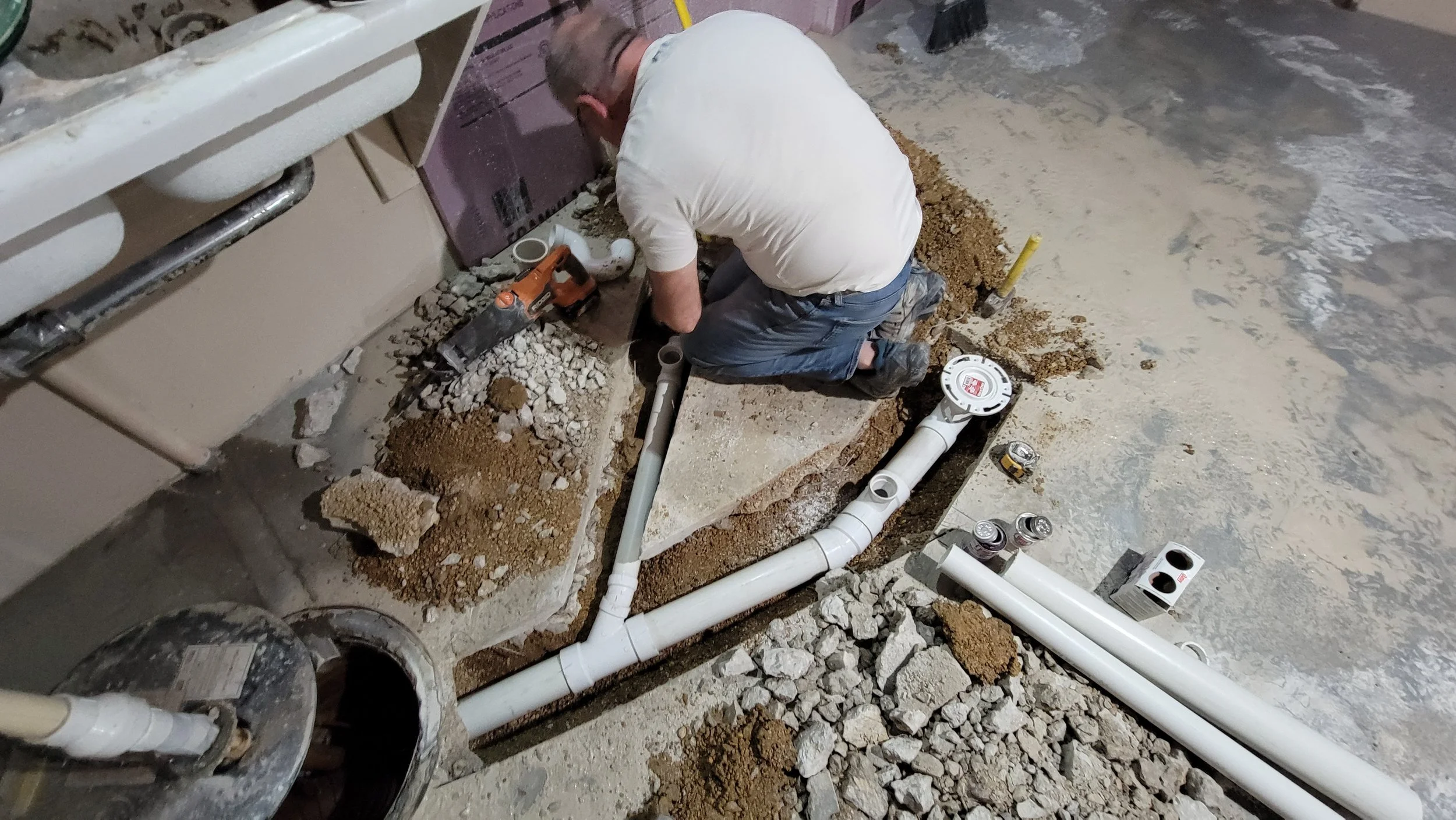 A man kneeling on the floor installing plumbing pipes under a sink, surrounded by construction tools, debris, and pipes on a concrete floor.