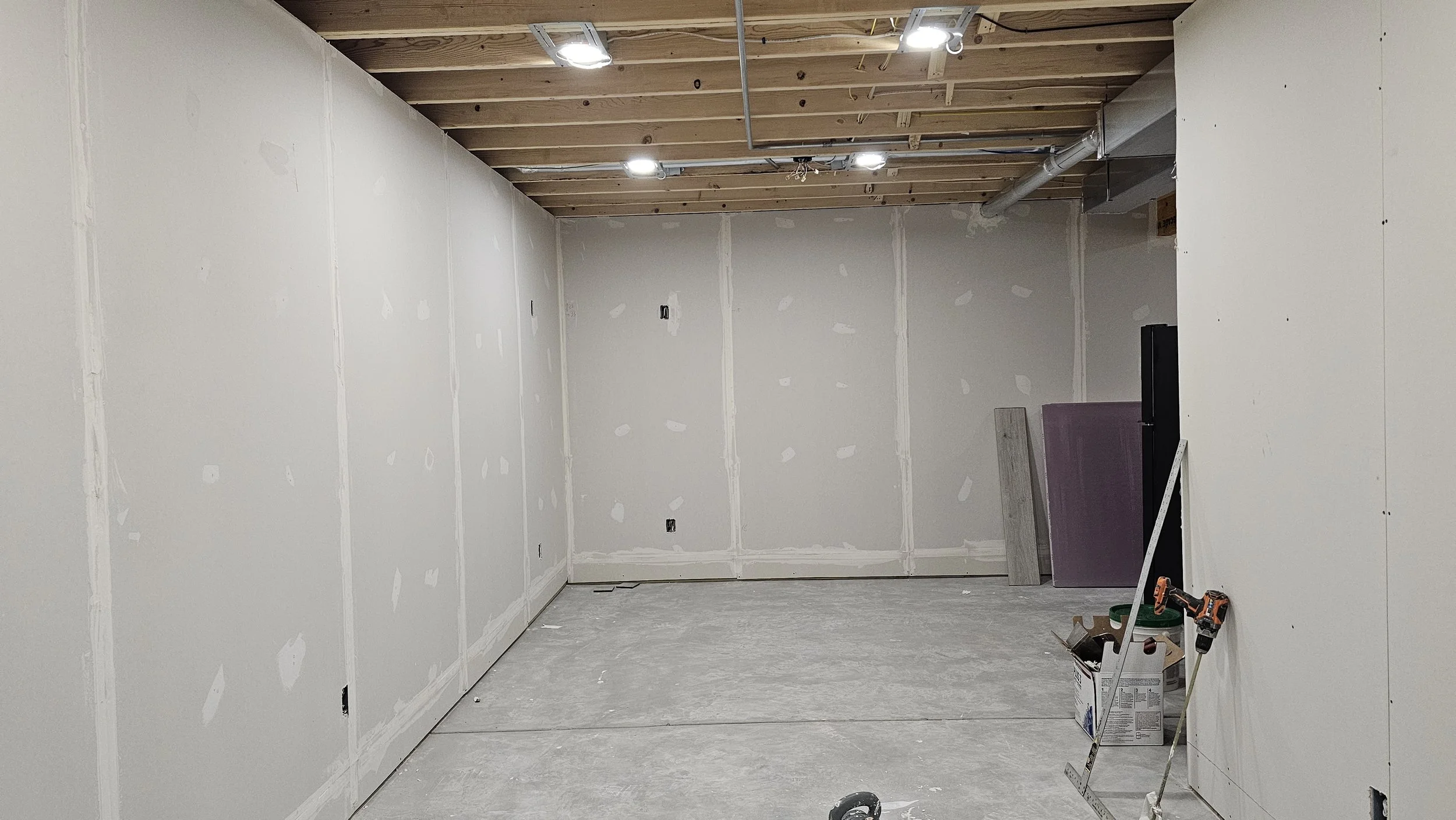 Interior of a room under construction with drywall installed, construction tools on the floor, and unfinished ceiling with exposed wood beams and electrical wiring.