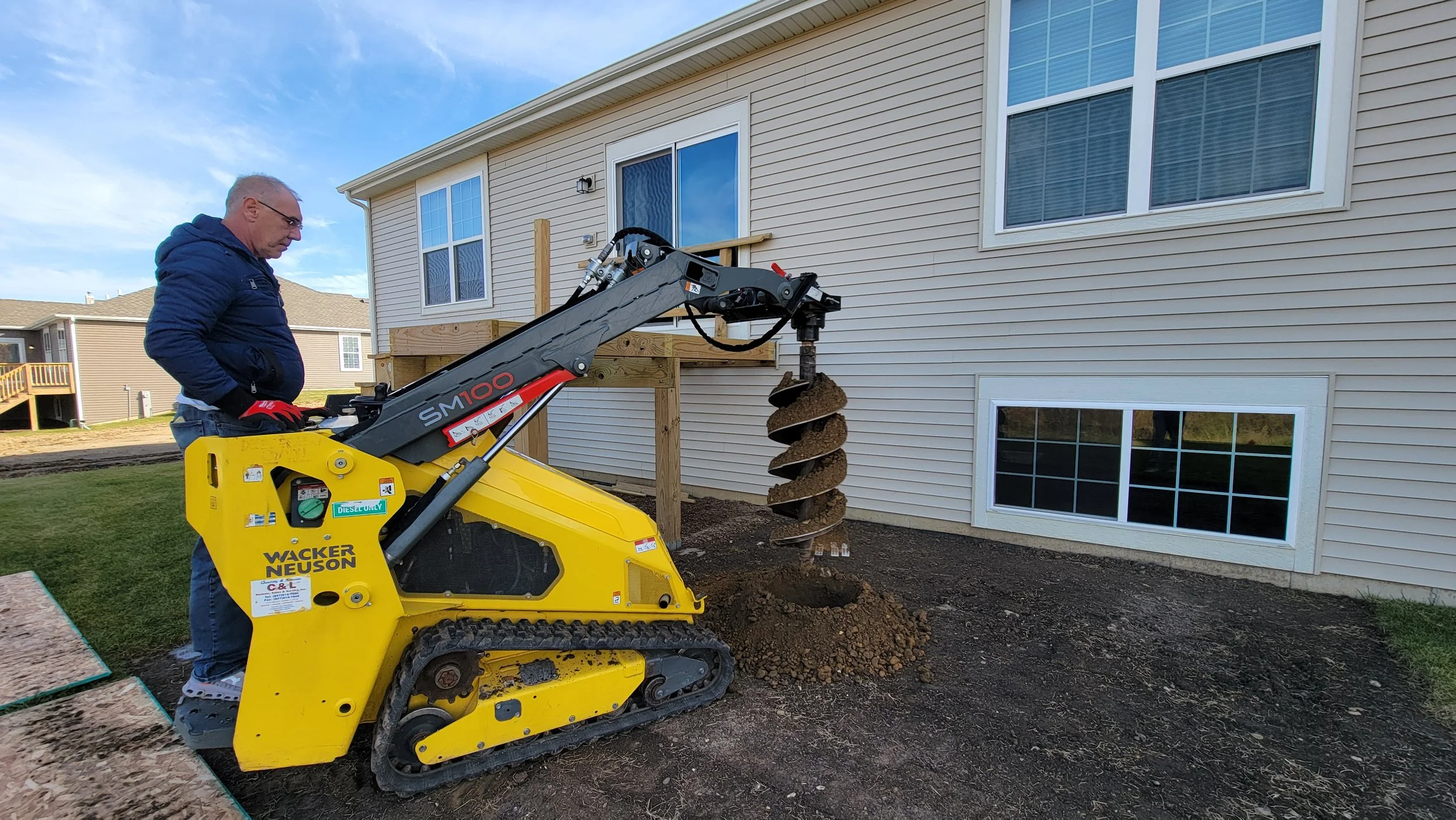 Man operating a small yellow Wacker Neuson compact excavator with a deep soil auger attachment, digging into the ground next to a beige house with white-framed windows.