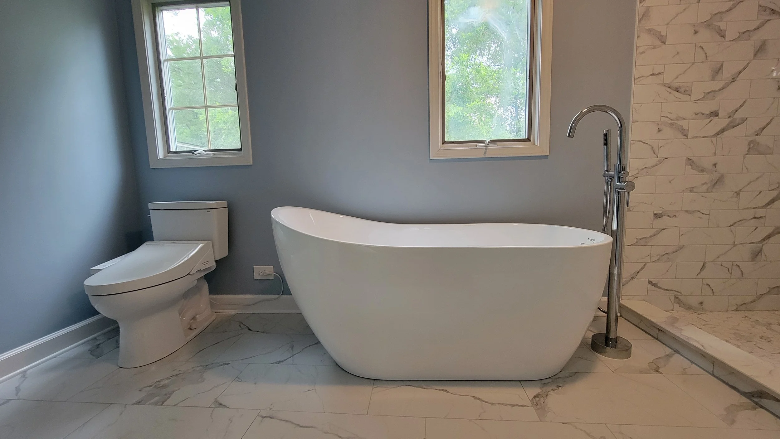 Modern bathroom with a white toilet, a freestanding bathtub near two windows, and a walk-in shower with marble tiles.