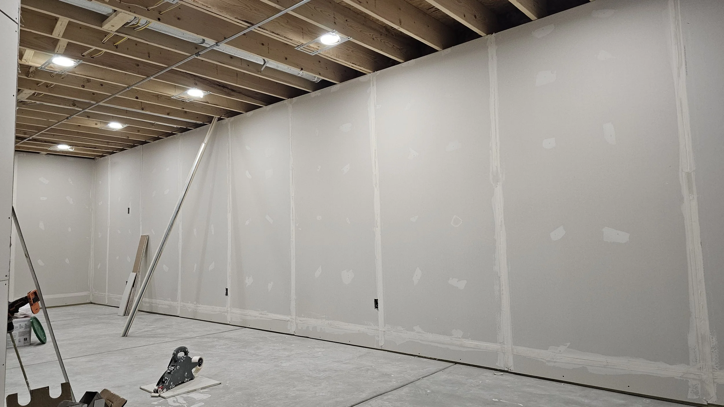 Interior of a room under construction with drywall walls, unfinished ceiling with exposed wooden beams, and construction tools and materials on the floor.