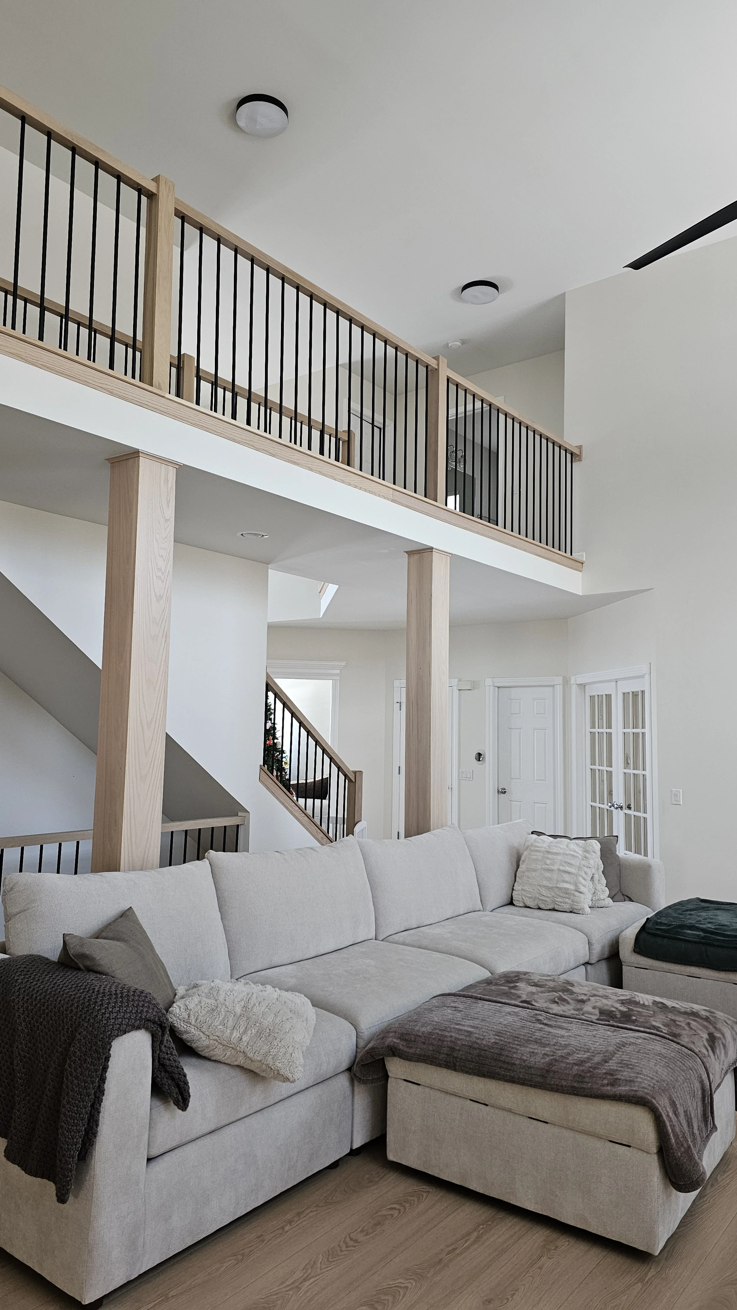 Living room with beige sectional sofa, throw pillows, gray blanket, wooden staircase with black railing, upstairs balcony with matching railing, white walls, wooden floors, front door in background.