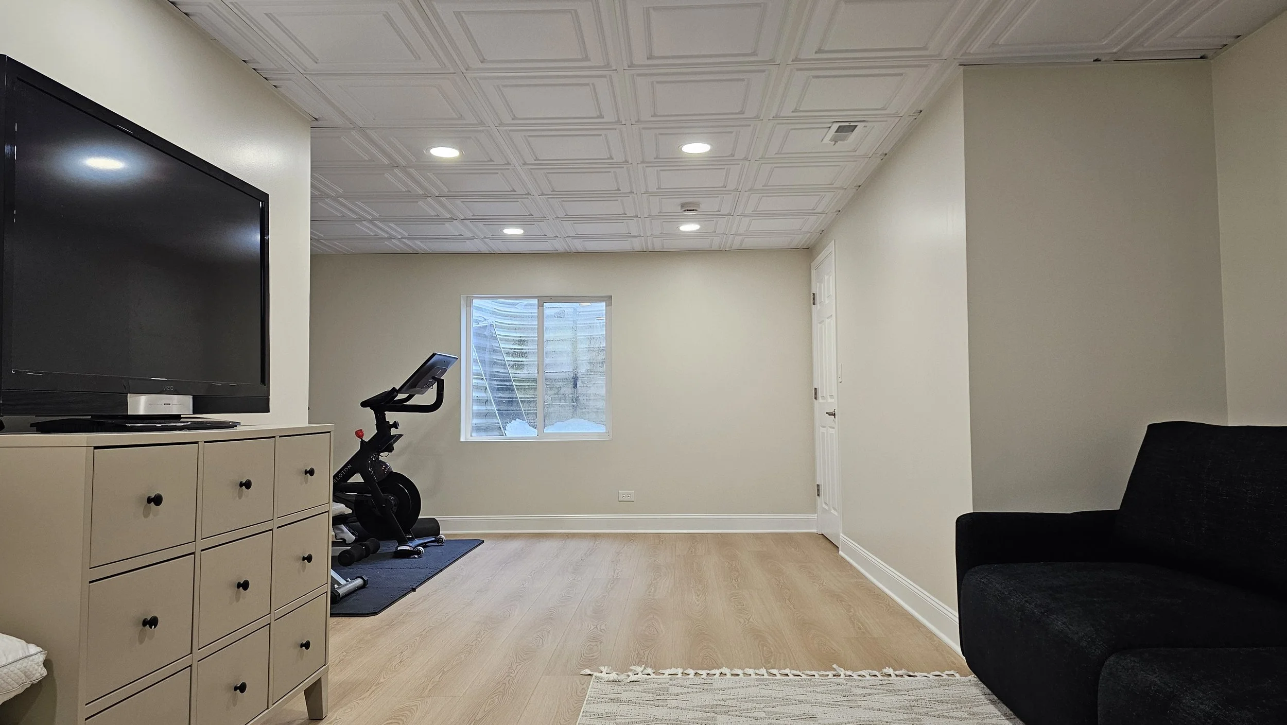 A living room with a TV on a beige storage cabinet, a black exercise bike, a window showing snow outside, a black couch on a light wood floor, and a beige rug.