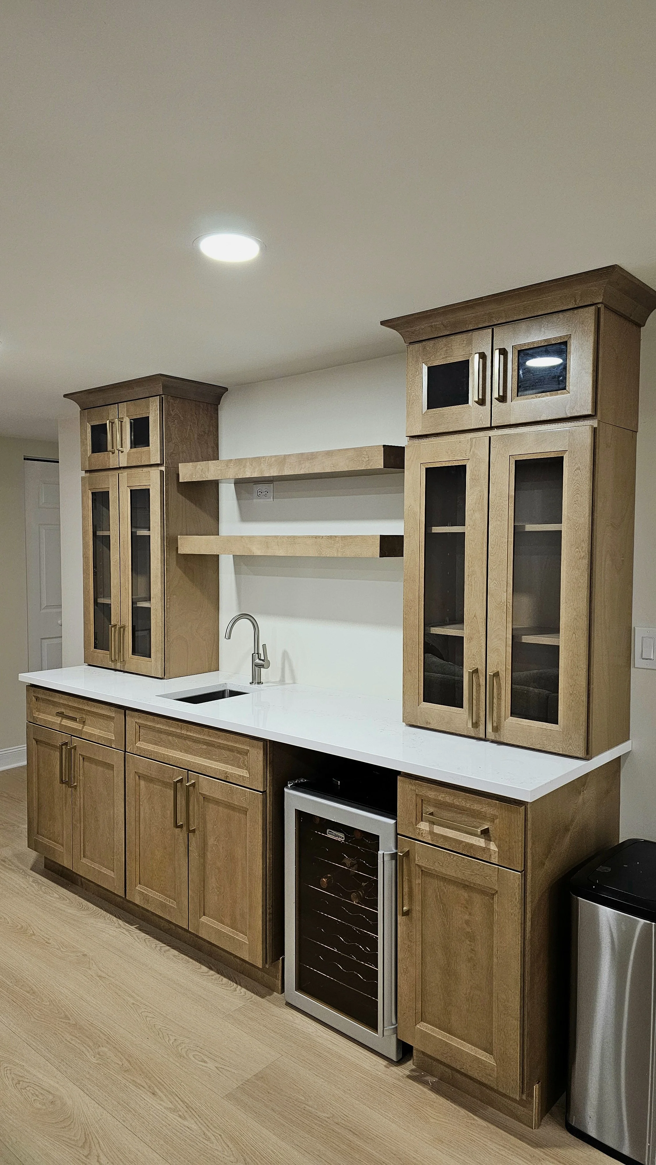 Kitchen with maple wood cabinets, white countertop, small wine cooler, and trash can, with open shelves above the sink and a ceiling light.